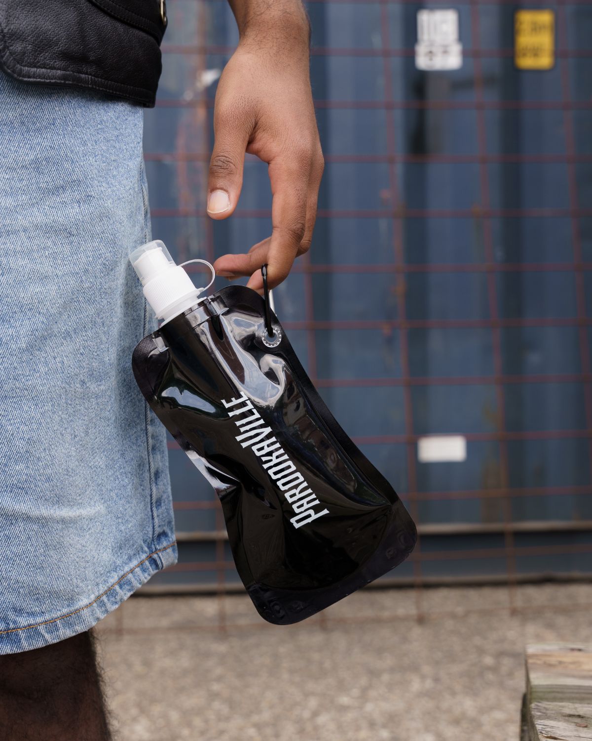 (DE) Ein Mann hält die schwarze faltbare Flasche mit einem Finger an dem an der Flasche befestigten Riemen fest. (EN) A black foldable bottle is dangling from a mans index finger that holds it at the string on the side. On the bottle there is the white inscription "PAROOKAVILLE".