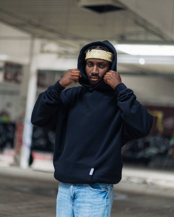 (DE) Ein Mann Er trägt einen schwarzen Hoodie und dazu eine Jeans. Außerdem trägt er eine gelbe Bandana mit braunem Muster. (EN) A man He is wearing a black hoodie and jeans. He is also wearing a yellow bandana with a brown pattern.