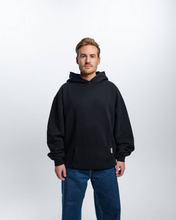 (DE) Ein Mann steht vor einem weißen Hintergrund. Er trägt einen schwarzen Hoodie und dazu eine Jeans. (EN) A man stands in front of a white background. He is wearing a black hoodie and jeans.