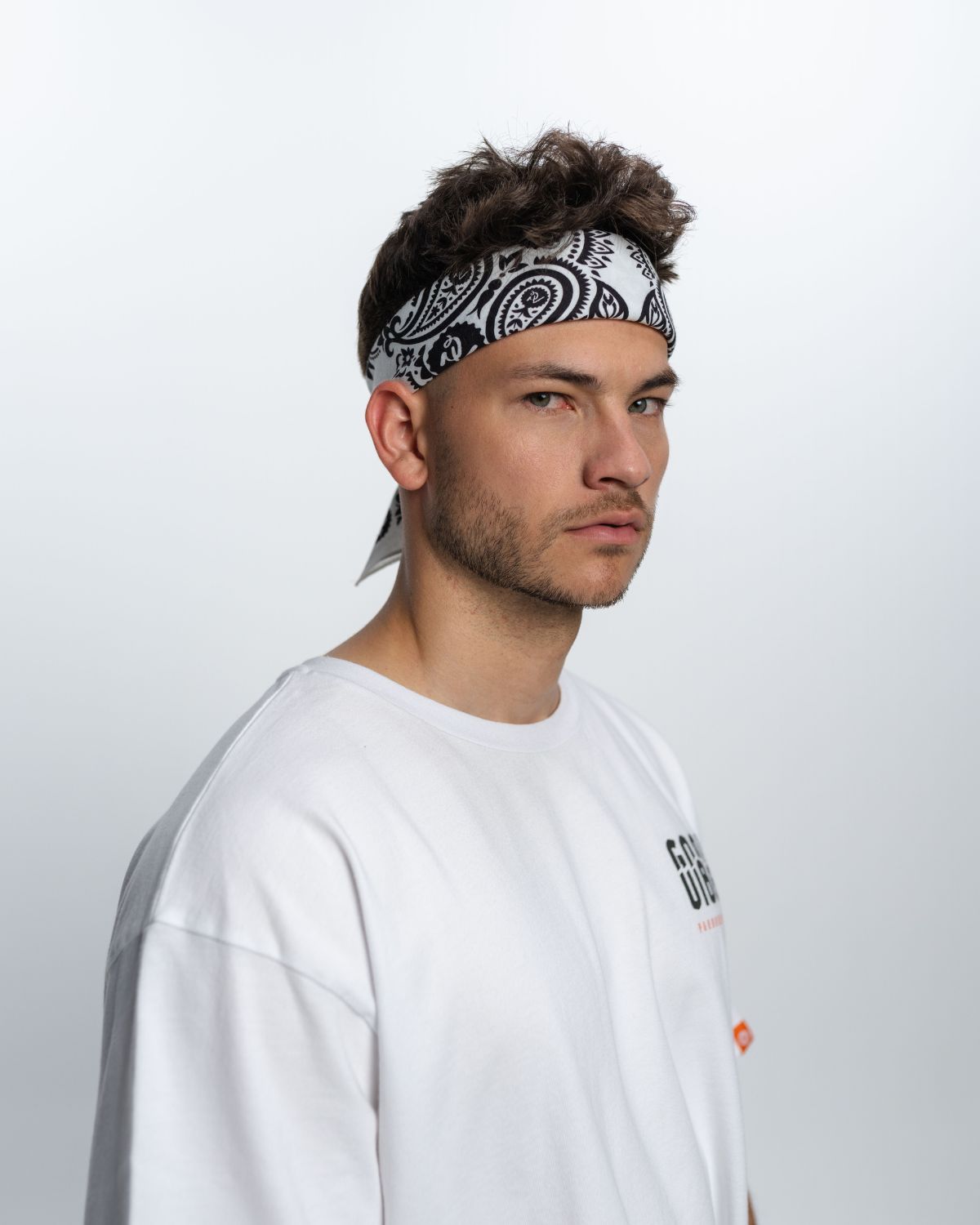 Ein Mann steht vor einem weißen Hintergrund. Er trägt ein weißes T-Shirt mit Aufschrift und eine schwarz weiße Bandana.