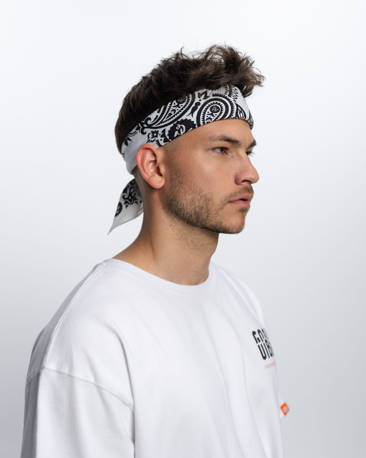 Ein Mann steht vor einem weißen Hintergrund. Er trägt ein weißes T-Shirt mit Aufschrift und eine schwarz weiße Bandana.