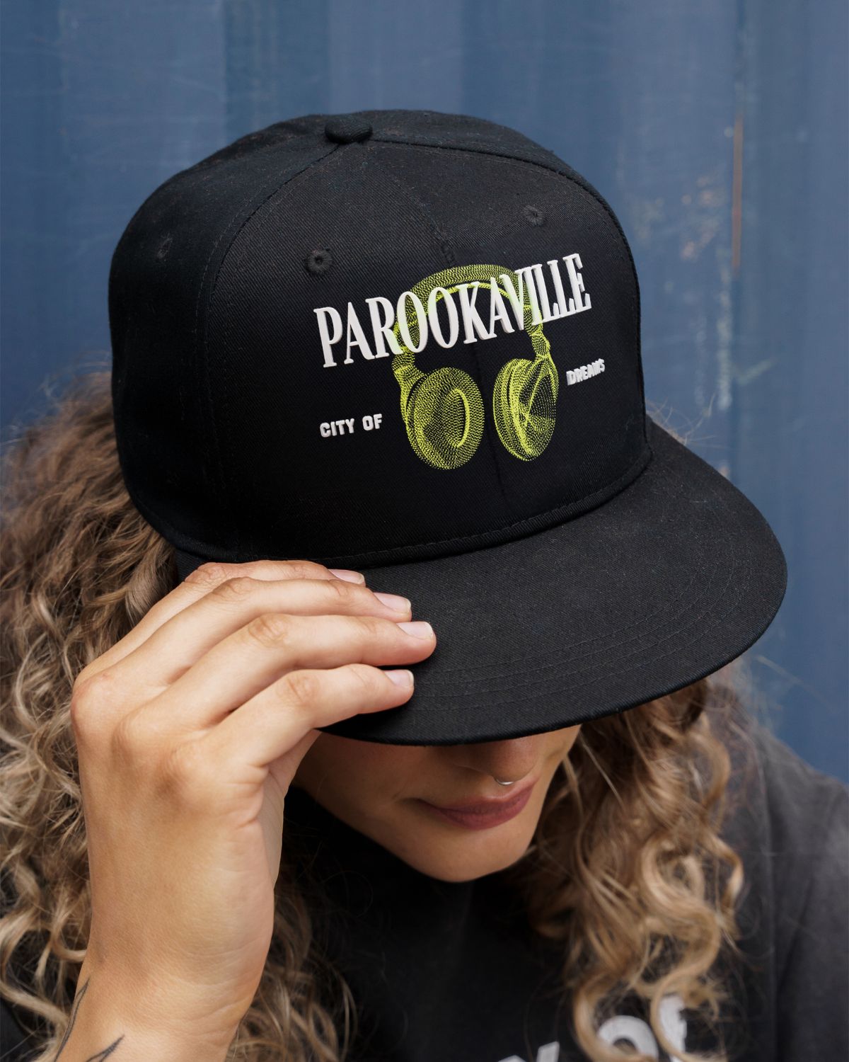 (DE) Eine Frau trägt ein dunkles T-Shirt mit weißer Aufschrift . Dazu trägt sie eine schwarze Cap mit der großen, weißen Aufschrift "PAROOKAVILLE" und der kleineren Unterschrift "CITY OF DREAMS" und einem Image von grünen headphones. (EN) A woman is wearing a dark T-shirt with white lettering. She is also wearing a black cap with the large, white inscription ‘PAROOKAVILLE’ and the smaller signature ‘CITY OF DREAMS’ and an image of green headphones.
