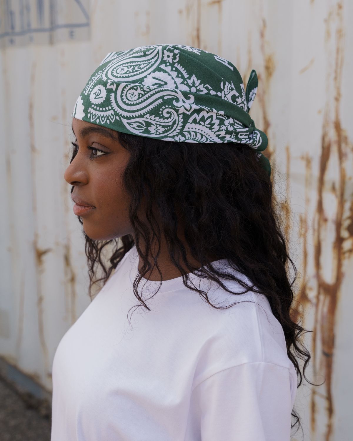 (DE) Eine Frau trägt ein weißes T-Shirt und dazu eine dunkelgrüne Bandana, welche sie um ihren Kopf gewickelt hat . Auf der Bandana sind weiße Blumenmuster zu erkennen. (EN) A woman is wearing a white T-shirt and a dark green bandana, which she has wrapped around her head. White floral patterns can be seen on the bandana.