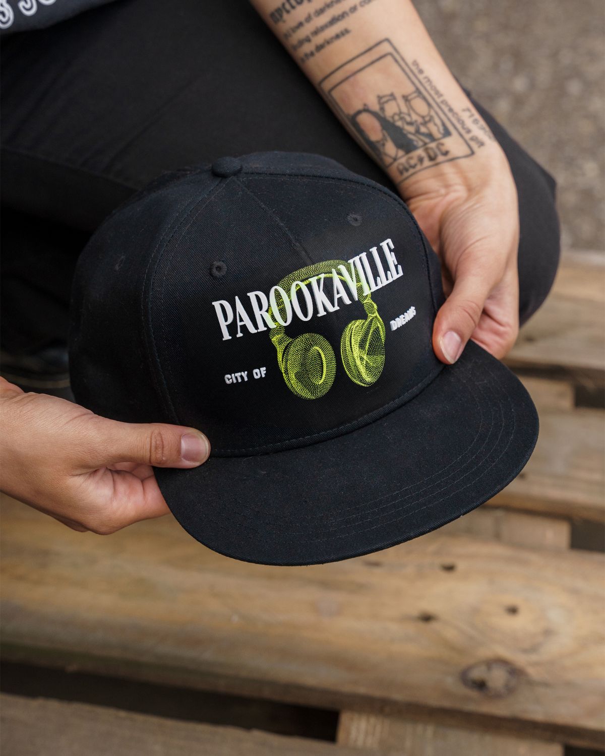 (DE) Eine Person hält eine schwarze Cap mit der großen, weißen Aufschrift "PAROOKAVILLE" und der kleineren Unterschrift "CITY OF DREAMS" und dem Image von grünen headphones in der Hand. (EN) One person is holding a black cap with the large, white inscription ‘PAROOKAVILLE’ and the smaller signature ‘CITY OF DREAMS’ and the image of green headphones.