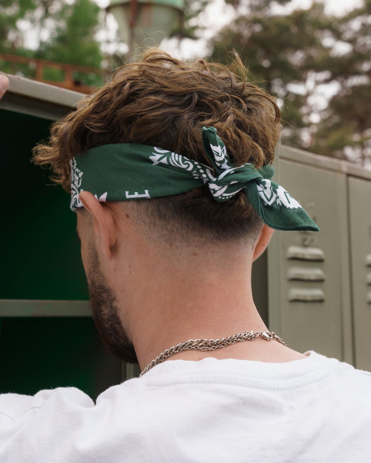 (DE) Ein Mann hat eine dunkelgrüne Bandana um seinen Kopf gewickelt und diese ist hinten zugeknotet. Auf der Bandana ist ein weißes Blumenmuster zu erkennen. (EN) A man has a dark green bandana wrapped around his head and it is knotted at the back. A white floral pattern can be seen on the bandana.