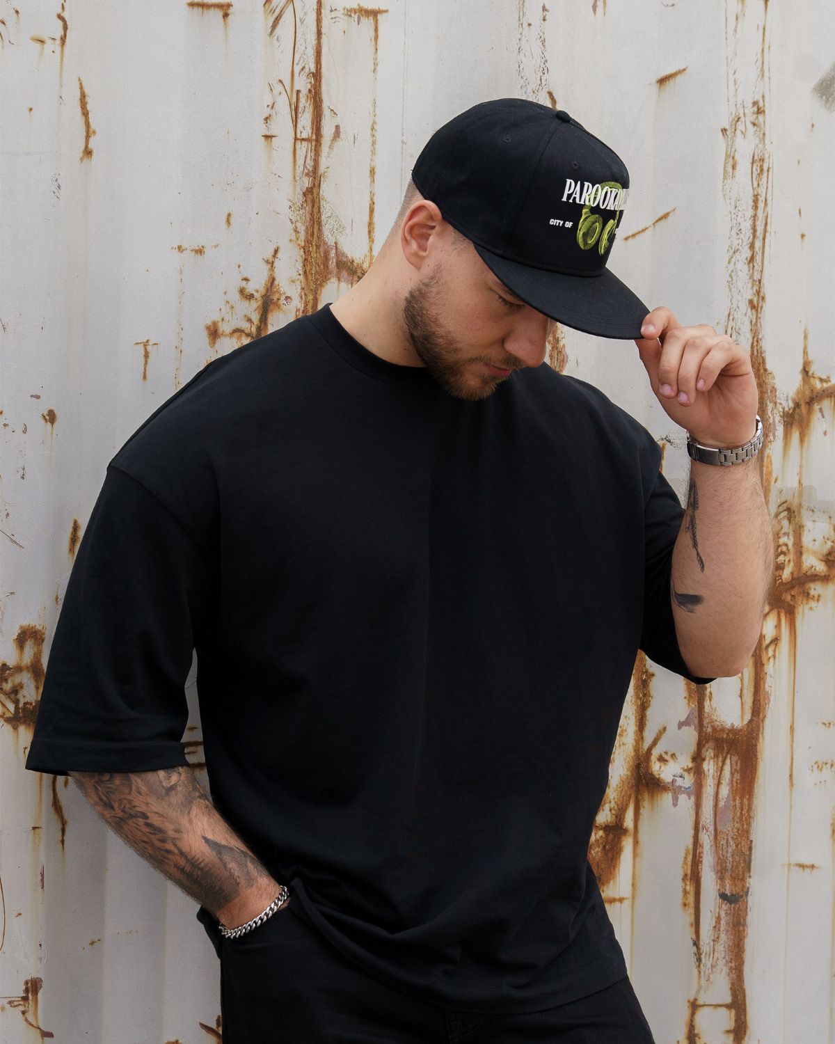 (DE) Ein Mann trägt ein schwarzes T-Shirt. Dazu trägt er eine schwarze Cap mit der großen, weißen Aufschrift "PAROOKAVILLE" und der kleineren Unterschrift "CITY OF DREAMS" und einem Image von grünen headphones. (EN) A man is wearing a black T-shirt. He is also wearing a black cap with the large, white inscription ‘PAROOKAVILLE’ and the smaller signature ‘CITY OF DREAMS’ and an image of green headphones.