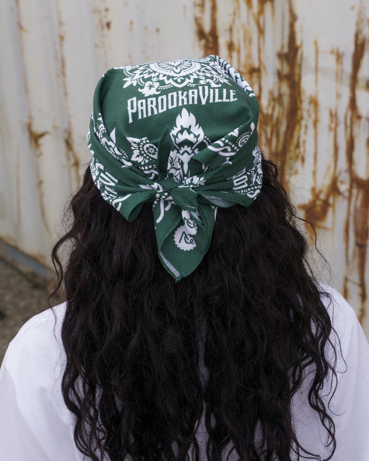 (DE) Eine Frau steht mit dem Rücken zur Kamera. Hinter ihr sind weiße, rostige Container. Dazu hat sie eine Bandana um ihren Kopf gewickelt. Die Bandana ist dunkelgrün und hat einen weißen PAROOKAVILLE Schriftzug. Außerdem ist dort noch ein Muster aus weißen Blumen zu erkennen. (EN) A woman stands with her back to the camera. Behind her are white, rusty containers. She has wrapped a bandana around her head. It is dark green and has white PAROOKAVILLE lettering. There is also a pattern of white flowers.