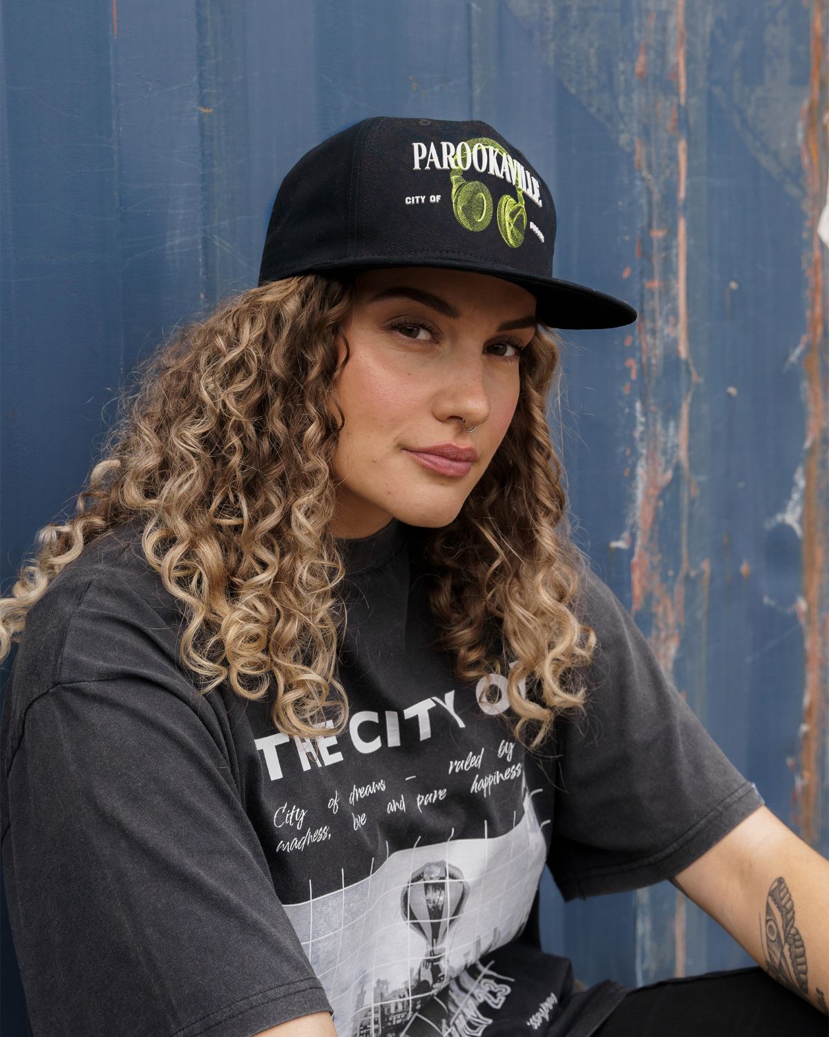 (DE) Eine Frau trägt ein dunkles T-Shirt mit weißer Aufschrift . Dazu trägt sie eine schwarze Cap mit der großen, weißen Aufschrift "PAROOKAVILLE" und der kleineren Unterschrift "CITY OF DREAMS" und einem Image von grünen headphones. (EN) A woman is wearing a dark T-shirt with white lettering. She is also wearing a black cap with the large, white inscription ‘PAROOKAVILLE’ and the smaller signature ‘CITY OF DREAMS’ and an image of green headphones.