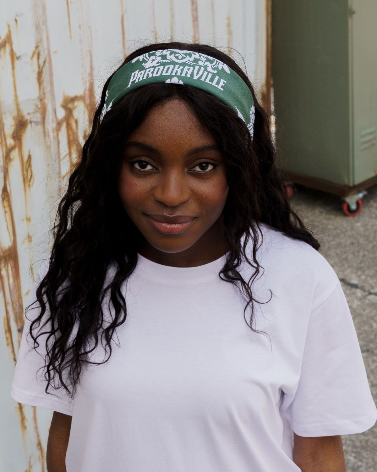 (DE) Eine Frau steht vor einem bunten Hintergrund aus Containern. Sie trägt ein weißes T-Shirt und dazu eine dunkelgrüne Bandana mit weißer Aufschrift "PAROOKAVILLE" und weißen Blumenprint. (EN) A woman stands in front of a colourful background of containers. She is wearing a white T-shirt and a dark green bandana with white lettering ‘PAROOKAVILLE’ and a white floral print.
