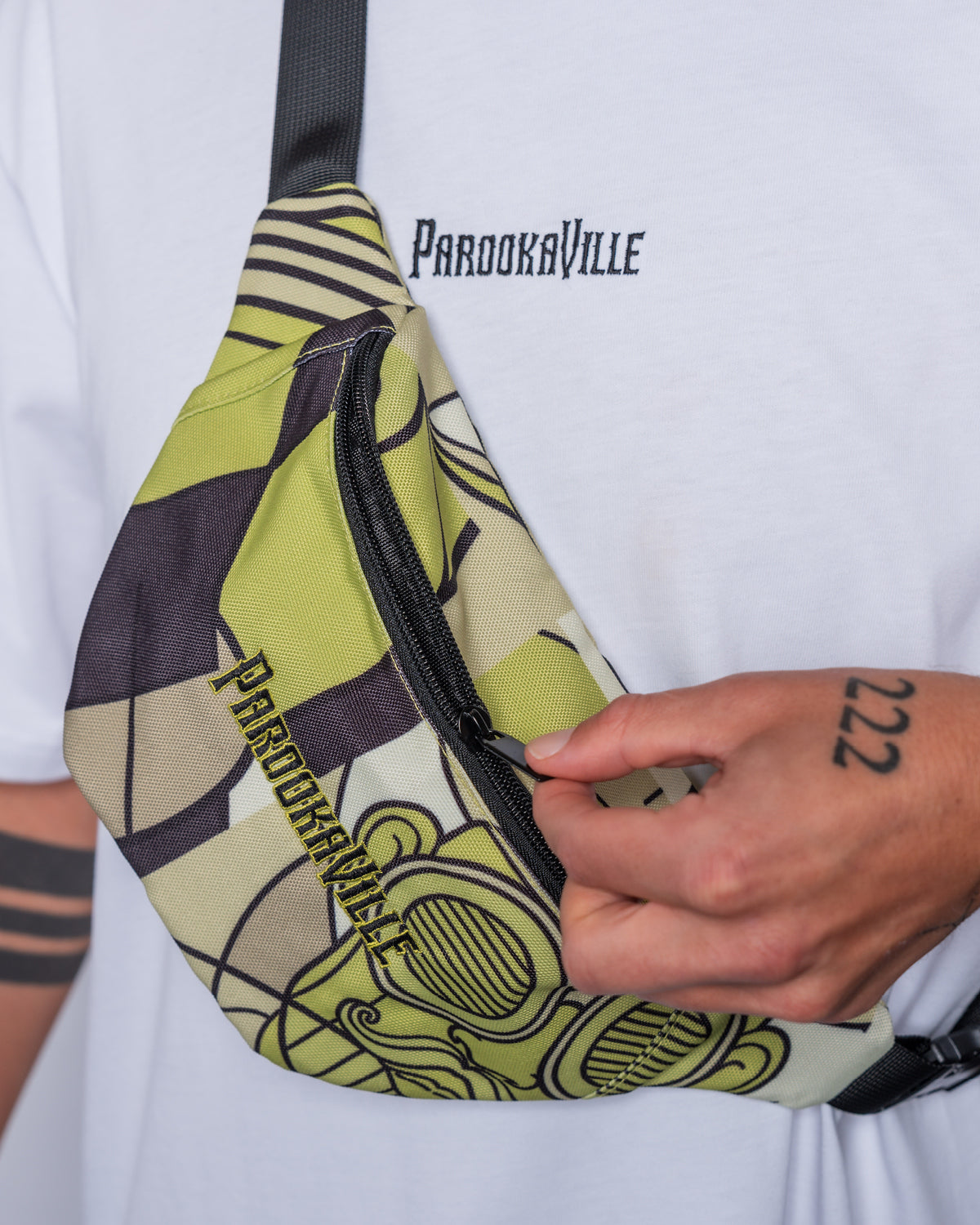 (DE) Ein Mann trägt die Bauchtasche mit weiß grünem Bill Print und dazu ein weißes T-Shirt mit dem schwarzen Schriftzug PAROOKAVILLE. (EN) A man is wearing a fanny pack with a Bill pattern in white and green. He combines the bag with a white T-Shirt with a black PV writing.