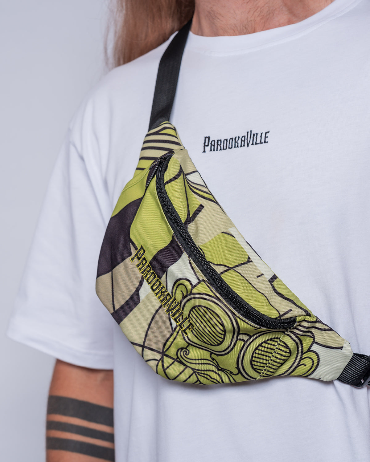 (DE) Ein Mann trägt die Bauchtasche mit weiß grünem Bill Print und dazu ein weißes T-Shirt mit dem schwarzen Schriftzug PAROOKAVILLE. (EN) A man is wearing a fanny pack with a Bill pattern in white and green. He combines the bag with a white T-Shirt with a black PV writing.