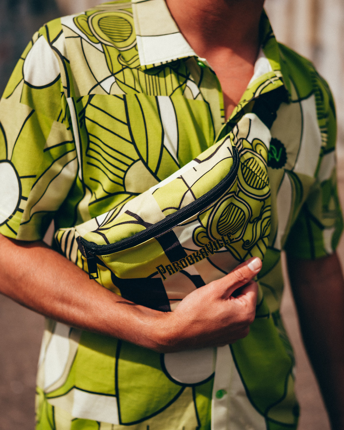 (DE) Ein Mann trägt die grün-weiß gemusterte BillPrint Bauchtasche. Die Tasche hat oben seitlich einen Reißverschluss und ist in einem hellgrün-weißen Billprint Motiv gemustert. Außerdem steht vorne auf der Tasche der schwarze Schriftzug PAROOKAVILLE. (EN) A man is wearing the PV Fanny Pack with Bill Print in a white and green pattern. There is a zip on the side at the top of the bag. Furthermore, the bag has the black inscription "PAROOKAVILLE" at the front.