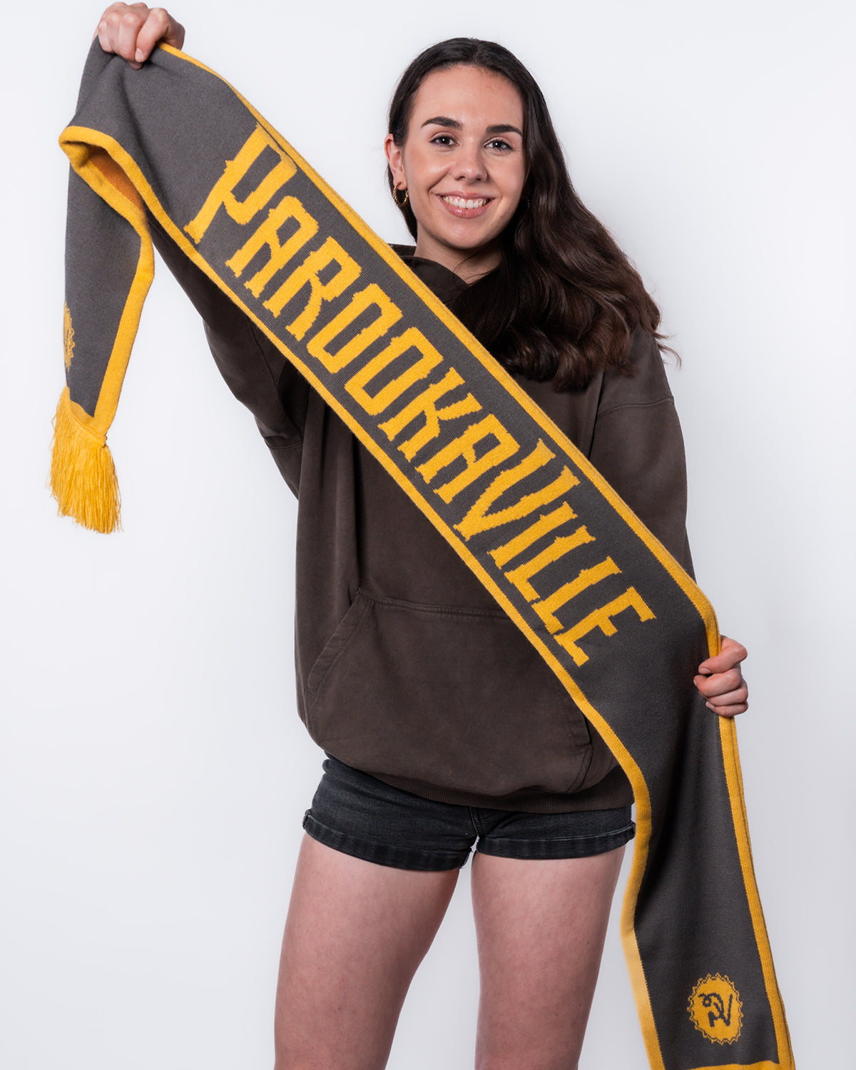 (DE) Eine Frau hält den PV Fan Schal mit der grauen Seite zur Kamera. Auf dem Schal steht in gelber Schrift "PAROOKAVILLE". (EN) A woman holds the grey side of the PV fan scarf in front of the camera. On the scarf is the bold lettering "PAROOKAVILLE".