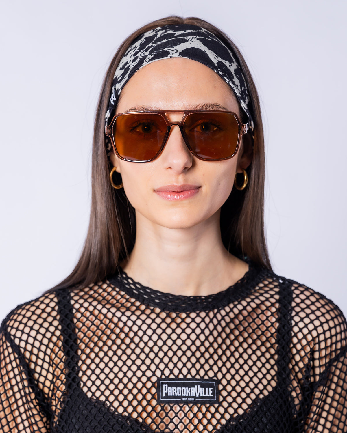 Eine Frau trägt das schwarz-weiße Haarband mit Leo-Muster in einer frontalen Studioaufnahme. Das Accessoire wird zu einer schwarzen Netzoberteil mit Parookaville-Patch und braun getönten Sonnenbrillen kombiniert.
