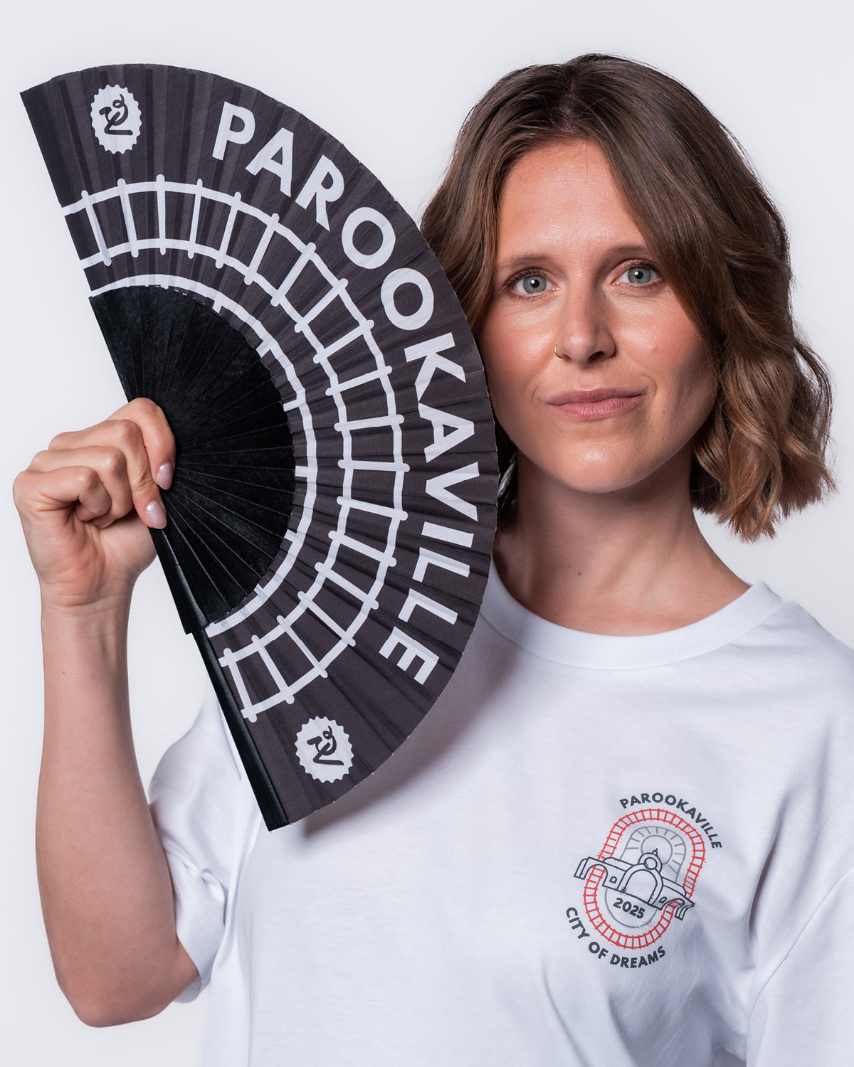 (DE) Eine Frau hält den Rails Fächer an ihr Gesicht. Der Fächer ist schwarz und hat ein Schienenmotiv in weiß aufgedruckt. Auch der PAROOKAVILLE Schriftzug sowie die PAROOKAVILLE Logos links und rechts sind weiß. (EN) A woman is holding the Rails hand fan with a rail pattern. The base is black and the pattern as well as the PAROOKAVILLE writing and the PV logos at the edges are white.