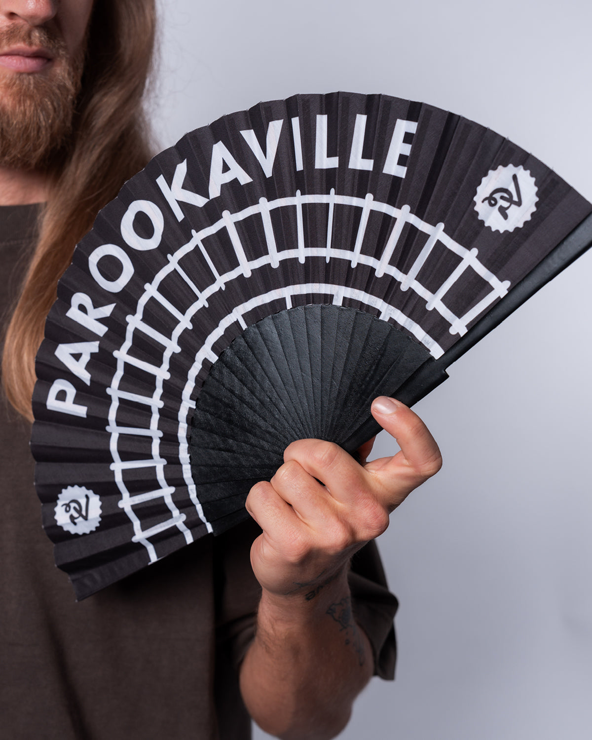 (DE) Ein Mann hält den PAROOKAVILLE Handfächer mit einem Schienenmuster in der Hand. Der Fächer selbst ist schwarz und das Schienenmotiv sowie der PAROOKAVILLE Schriftzug darüber ist weiß. (EN) A man is holding the PAROOKAVILLE Rails hand fan with a rail pattern. The base is black and the pattern as well as the PAROOKAVILLE writing are white.