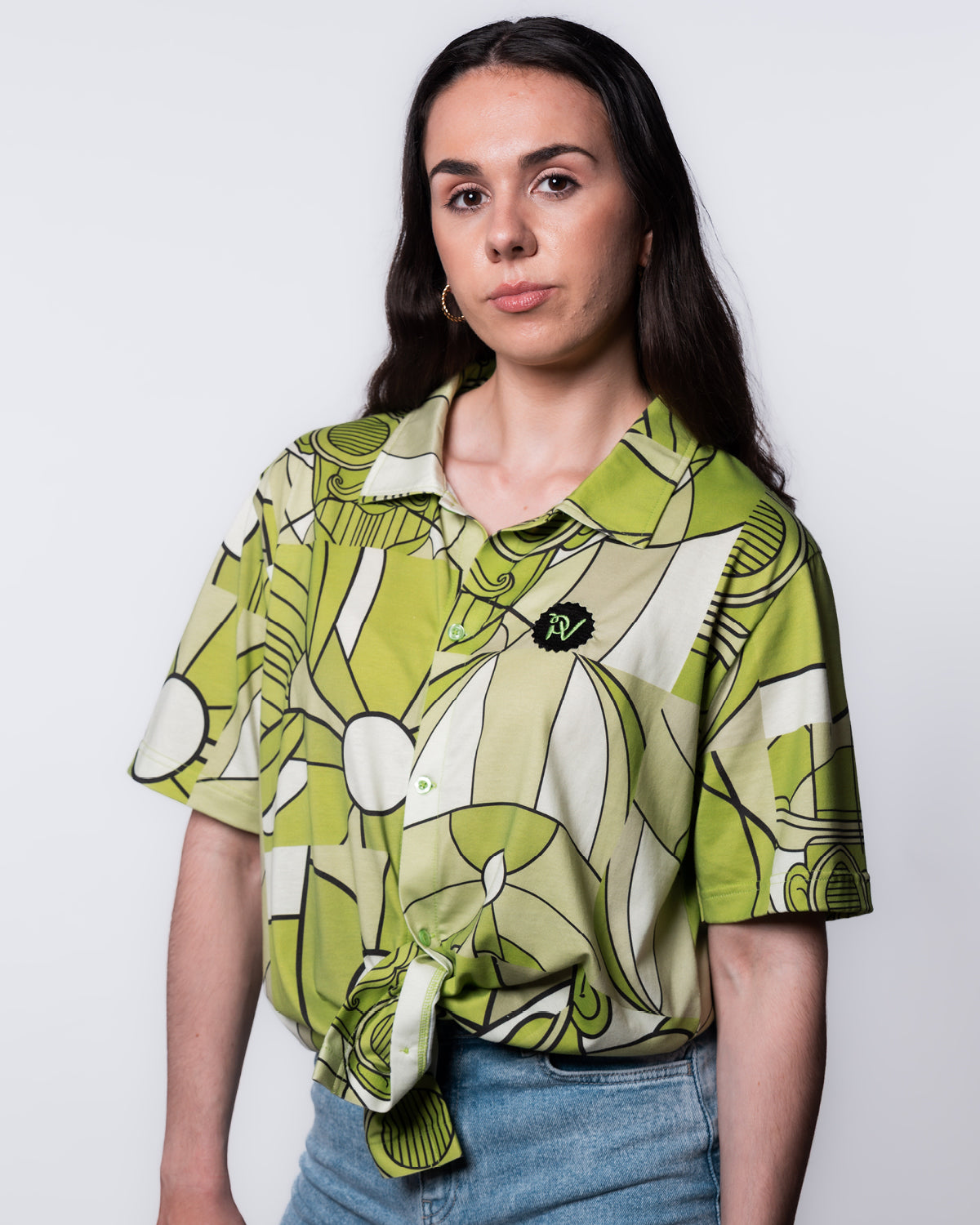 (DE) Eine Frau trägt das Hemd mit weiß-grünem Bill Print mit Motiven von Bill Parooka in hellgrün. Außerdem hat das Hemd recht oben das PAROOKAVILLE Logo in grün auf schwarzem Untergrund aufgestickt. (EN) A woman is wearing a shirt with Bill Print in white and green. There is a green PV logo embroided on a black base.