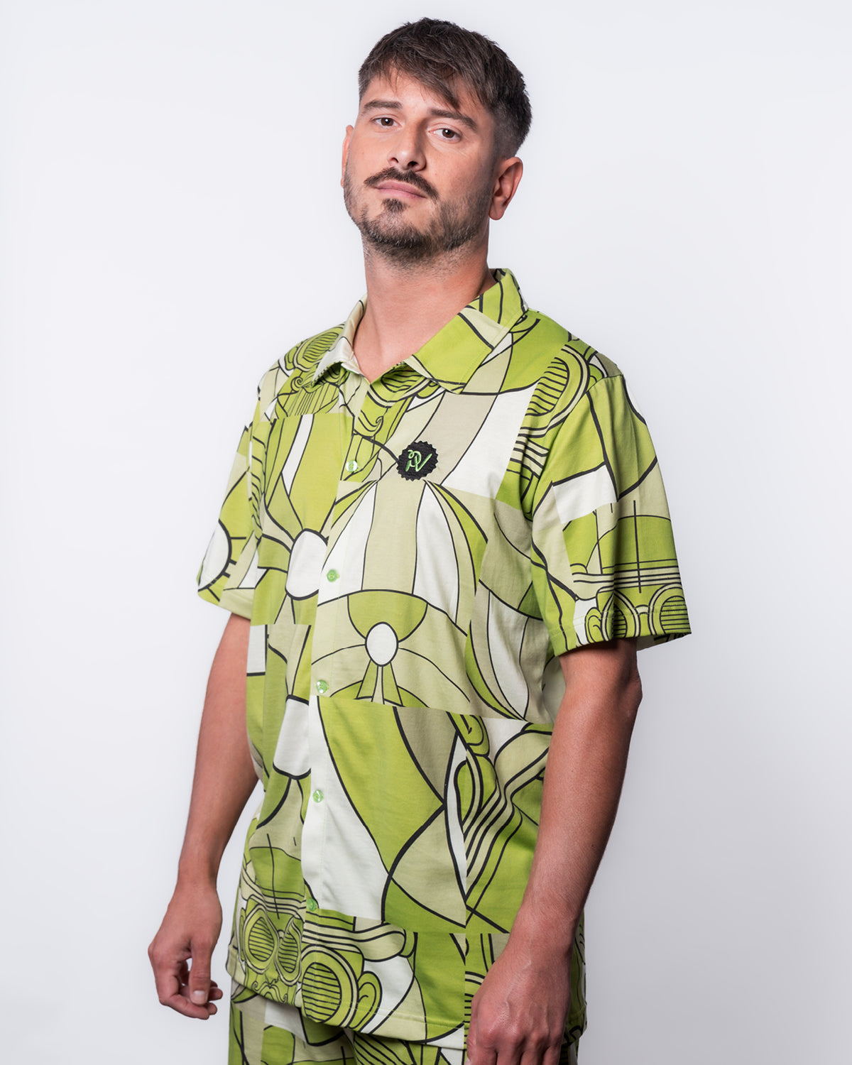 (DE) Ein Mann trägt das Hemd mit weiß-grünem Bill-Print und dazu die passende Shorts. Rechts oben ist das PV Logo in grün auf schwarzem Untergrund zu sehen. (EN) A man is wearing a shirt with Bill Print in white and green. At the right top corner at the front there is a green PV emblem embroided on a black base.