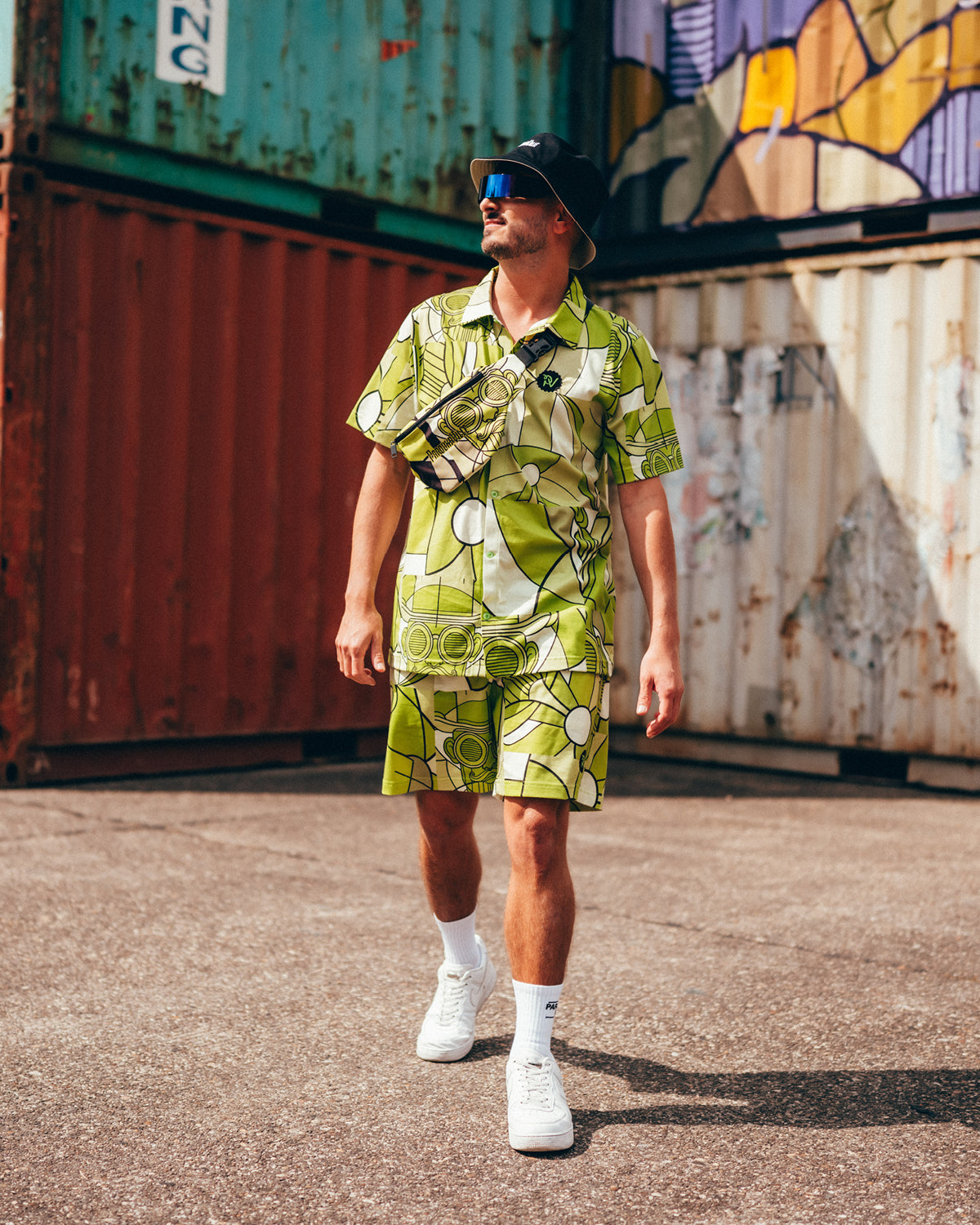 (DE) Ein Mann steht vor bunten Containern. Er trägt ein Hemd mit einem grafischen all-over Print. Auf dem Print ist auch eine Zeichnung von Bill Parooka zu sehen. Die base ist weiß und der Print ist mehrfarbig grün. Passend zum Hemd trägt er eine Shorts. (EN) A man stands in front of colourful containers. He is wearing a shirt with an all-over graphic print. The print also features a drawing by Bill Parooka. The base is white and the print is multicoloured green. He is wearing shorts to match the shirt.