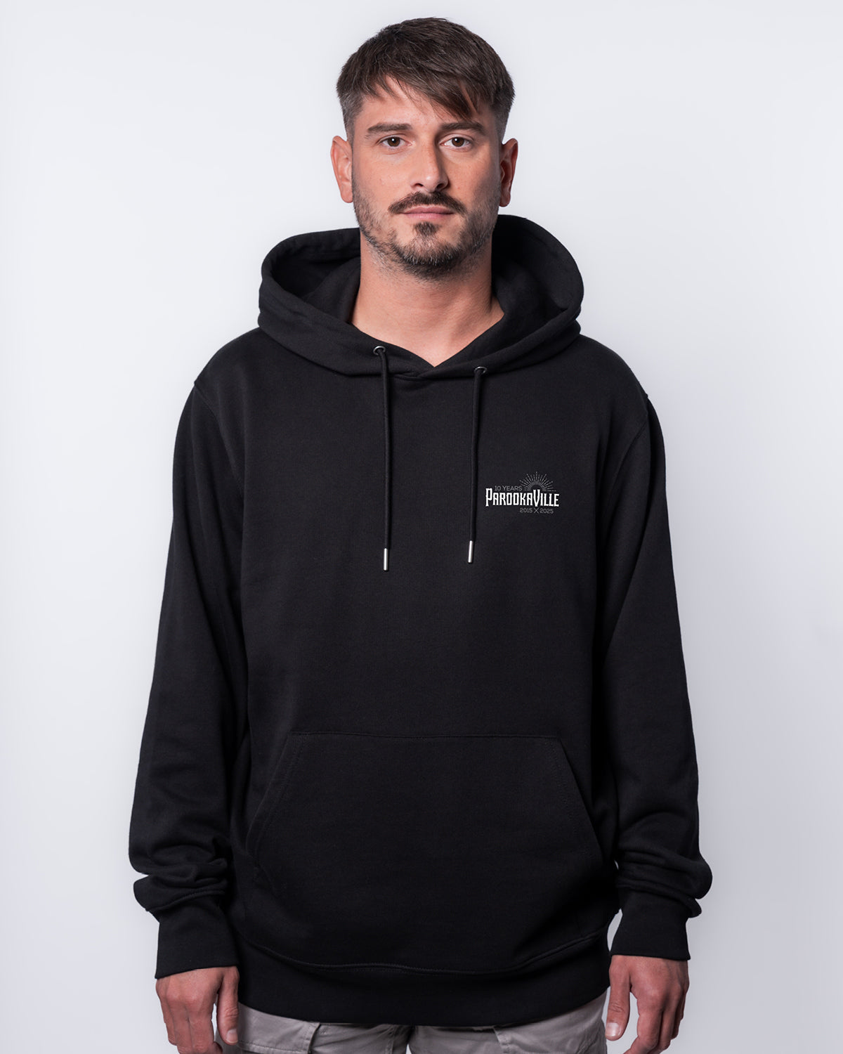 (DE) Ein Mann trägt einen schwarzen Hoodie mit dem weißen 10 Years Parookaville- Logo auf der rechten Oberseite. Außerdem sind an den Enden der Kapuze Kordeln, um diese festzubinden. (EN) A man is wearing a black hoodie with the white 10 Years Parookaville logo on the top right. There are also cords at the ends of the hood to tie it up.