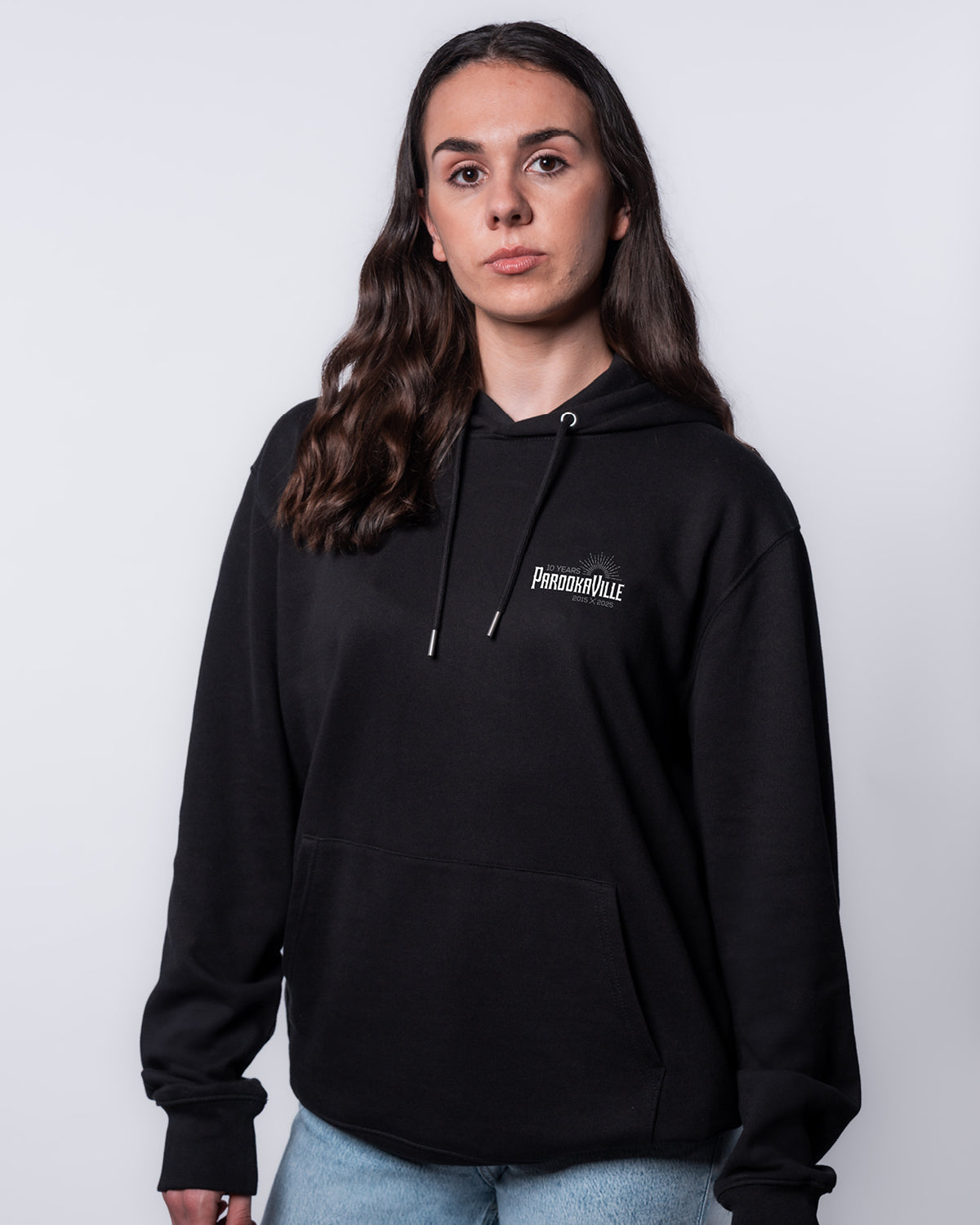 (DE) Eine Frau trägt den schwarzen Line-Up Hoodie mit einem weißen 10 Years PAROOKAVILLE Schriftzug auf der rechten Oberseite. Zusätzlich hat er vorne eine große Bauchtasche. An den Enden der Kapuze sind Kordeln, um diese zu befestigen. (EN) A woman wears the black line-up hoodie with white 10 Years PAROOKAVILLE lettering on the top right. It also has a large belly pocket at the front. There are cords at the ends of the hood to fasten it.