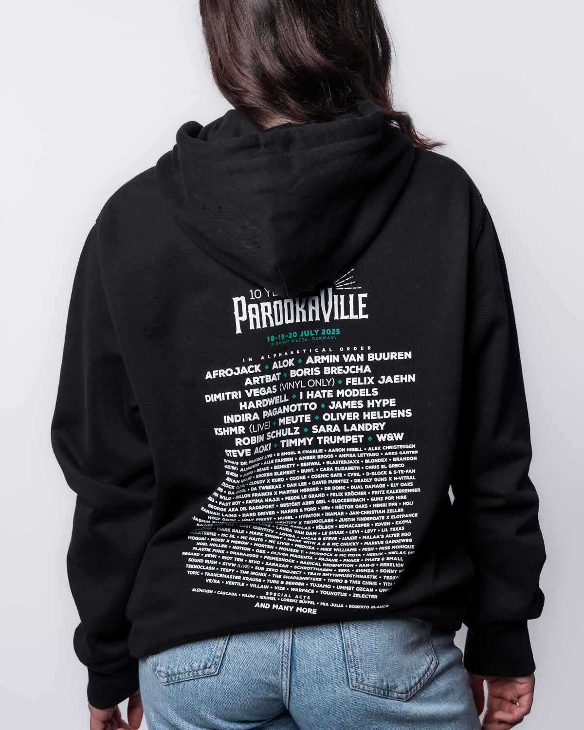 (DE) Eine Frau trägt den schwarzen Line-Up Hoodie mit weißer Schrift auf dem Back Print. Auf der Rückseite stehen die Artists unter der 10 Years PAROOKAVILLE Aufschrift. (EN) A woman wears the black line-up hoodie with white lettering on the back print. On the back are the artists under the 10 Years PAROOKAVILLE inscription.