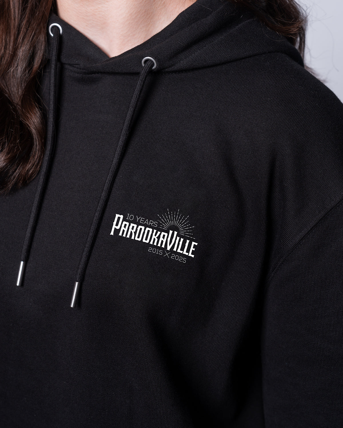 (DE) Man sieht eine Nahansicht des schwarzen Line-Up Hoodies mit dem 10 Years PAROOKAVILLE Schriftzug auf der rechten Oberseite.Außerdem sind an den Enden der Kapuze Kordeln, um diese zu befestigen. (EN) You can see a close-up view of the black line-up hoodie with the 10 Years PAROOKAVILLE lettering on the top right, and there are also cords at the ends of the hood to fasten it.