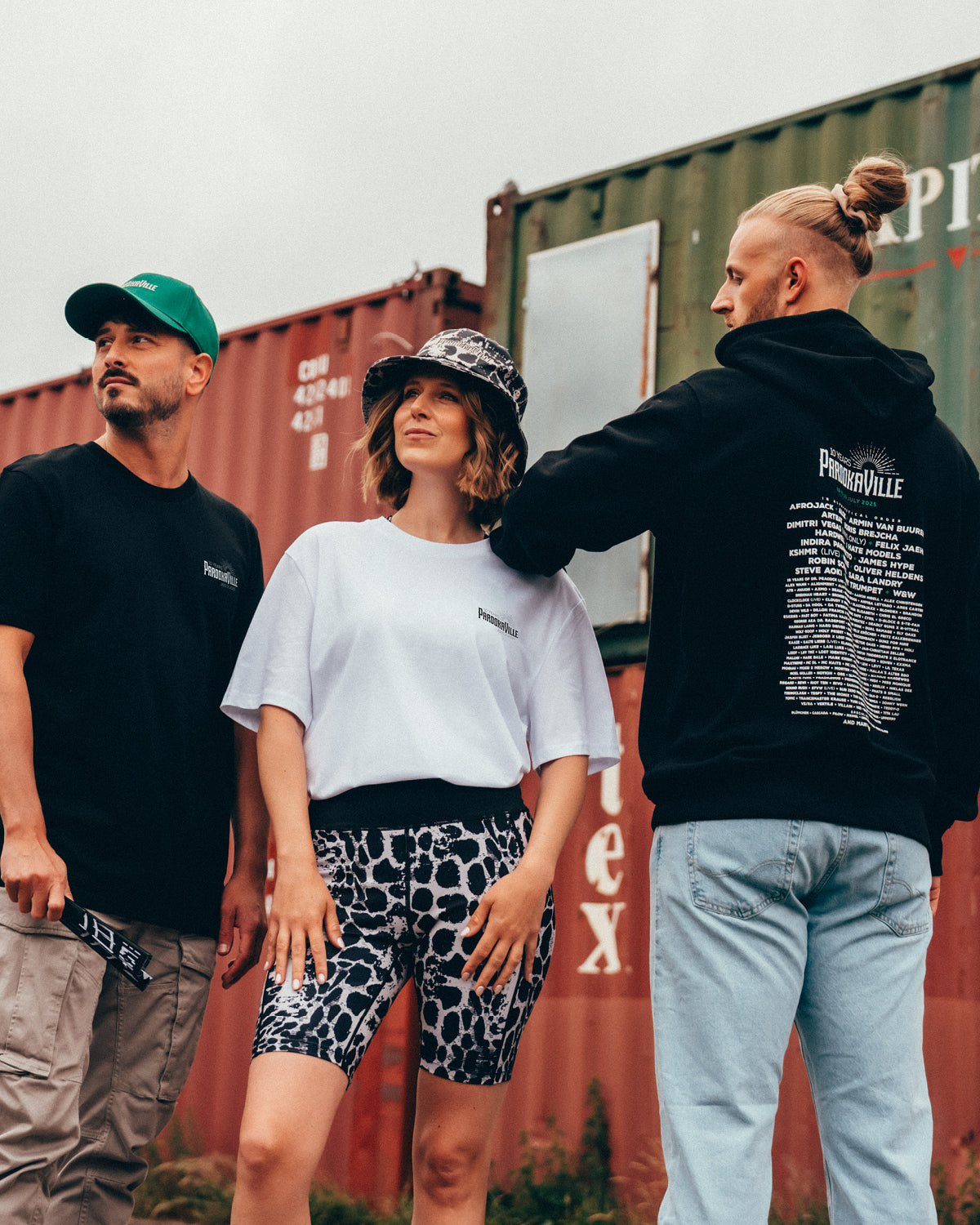 (DE) Drei Personen stehen vor bunten Containern. Die beiden Männer tragen schwarze Überteile, auf der Rückseite vom Hoodie des Mannes rechts ist das Line-Up 2025 zu erkennen. Die Frau trägt ein Sportoutfit mit Leopardenprint und dazu einen Bucket Hat. (EN) Three people stand in front of colourful containers. The two men are wearing black tops and the line-up 2025 can be seen on the back of the hoodie worn by the man on the right. The woman is wearing a sports outfit with a leopard print and a bucket hat.