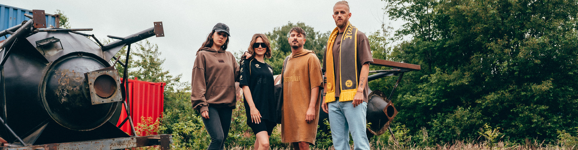 (DE) Zwei Personen tragen Oberteile in einem Braunton, die Frau mittig trägt das schwarze PAROOKAVILLE- Trikot und ein Mann trägt einen braunen Poncho. Außerdem trägt der Mann rechts rau-goldenen Jubiläumsschal. (EN) Two people are wearing tops in a shade of brown, the woman in the centre is wearing the black PAROOKAVILLE jersey and one man is wearing a brown poncho. The man on the right is also wearing a rough gold anniversary scarf.