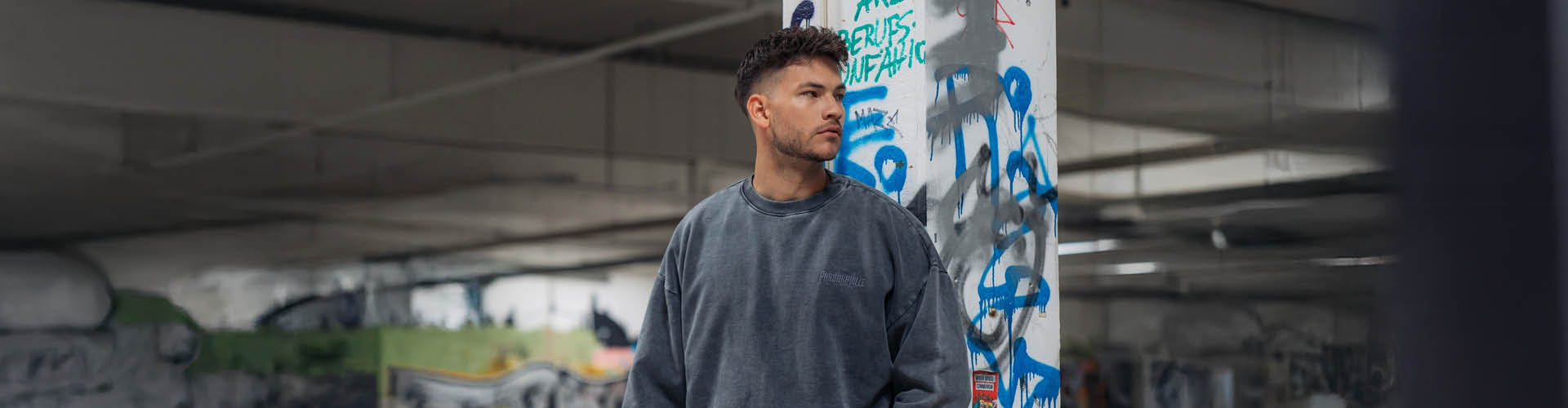 (DE) Ein Mann steht vor einem bunten Graffiti Hintergrund. Er trägt einen dunkelgrauen Pullover mit der dunklen Aufschrift "PAROOKAVILLE" rechts oben. (EN) A man is standing in front of a colourful graffiti background . He is wearing a dark sweater with the dark inscription "PAROOKAVILLE" at the top right front corner.