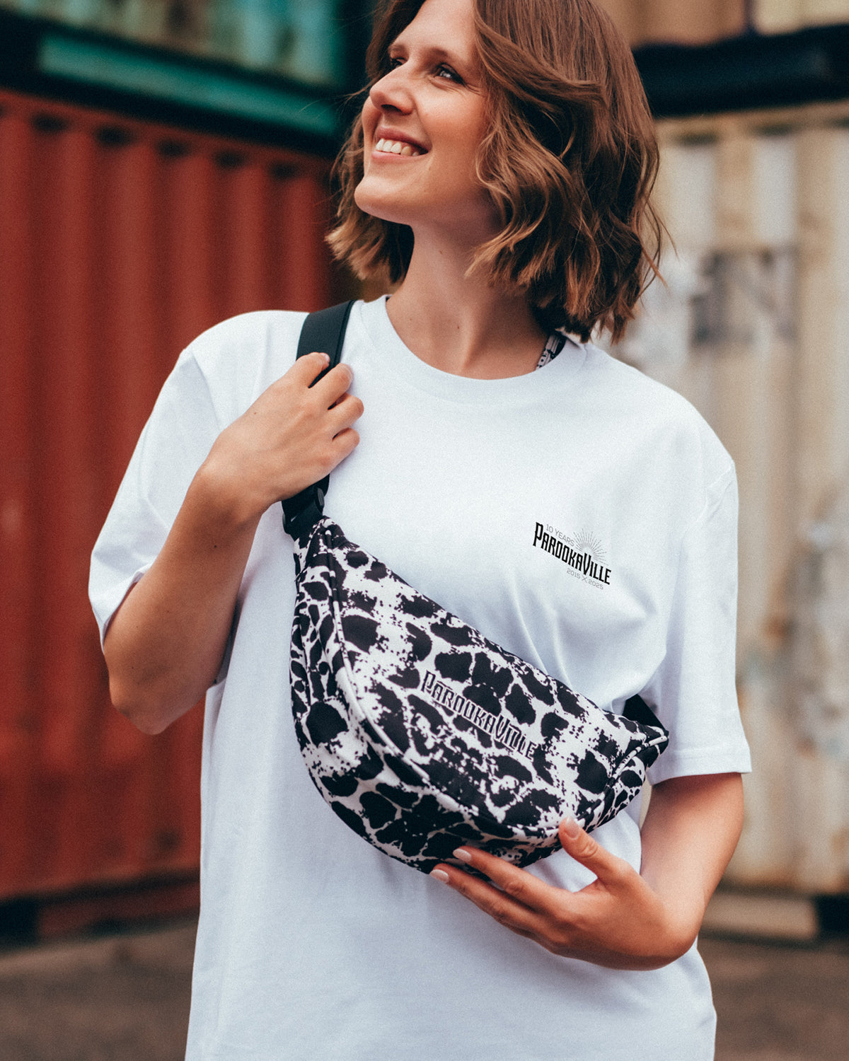 (DE) Eine Frau steht vor bunten Containern. Sie trägt die schwarz-weiß gefleckte Moonbag-Tasche im Leopardenmuster und dazu ein weißes PAROOKAVILLE T-Shirt mit einem schwarzen 10Years PAROOKAVILLE-Logo auf der linken Oberseite. (EN) A woman stands in front of colourful containers. She is carrying the black and white spotted Moonbag bag in leopard print and a white PAROOKAVILLE T-shirt with a black 10Years PAROOKAVILLE logo on the top left.