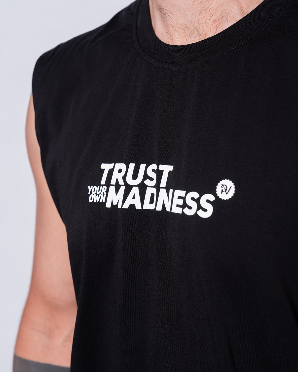 (DE) Man sieht eine Nahansicht des schwarzen, ärmellosen Muskelshirts "Madness" mit der weißen Aufschrift "TRUST YOUR OWN MADNESS" mittig auf der Vorderseite. (EN) You can see a close-up of the black, sleeveless muscle shirt ‘Madness’ with the white inscription ‘TRUST YOUR OWN MADNESS’ in the centre of the front.