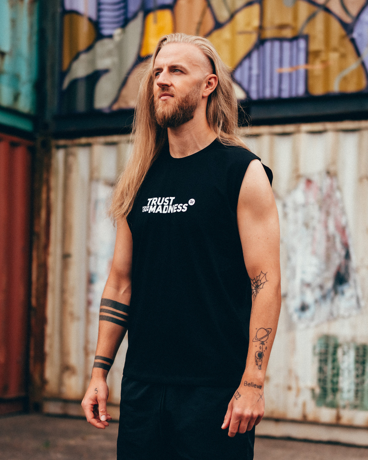 (DE) Ein Mann trägt ein ärmelloses schwarzes Shirt mit dem weißen Schriftzug "TRUST YOUR OWN MADNESS" und dem PAROOKAVILLE Emblem. (EN) A man wears a sleeveless black shirt with the white lettering ‘TRUST YOUR OWN MADNESS’ and the PAROOKAVILLE emblem.