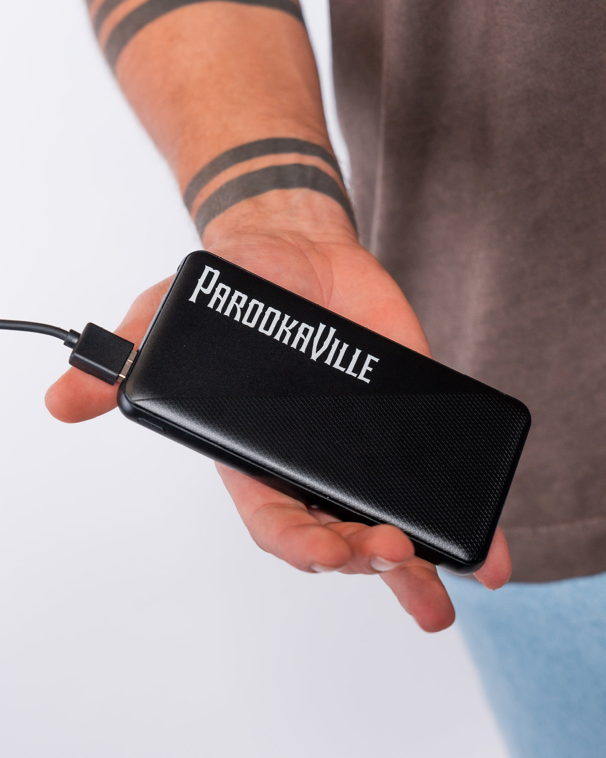 (DE) Eine Person hält die schwarze Powerbank mit dem weißen Schriftzug "PAROOKAVILLE" in die Kamera. (EN) A person holds the black powerbank with the white lettering ‘PAROOKAVILLE’ up to the camera.