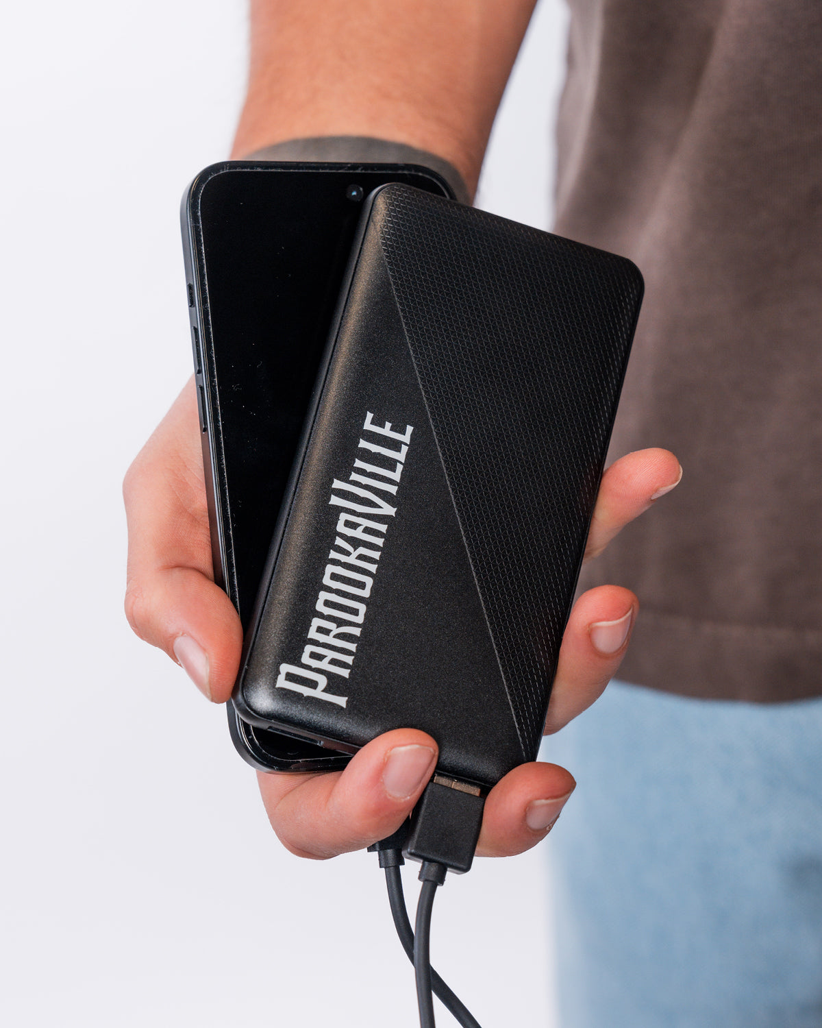 (DE) Eine Person hält die schwarze Powerbank mit dem weißen Schriftzug "PAROOKAVILLE" in die Kamera. (EN) A person holds the black powerbank with the white lettering ‘PAROOKAVILLE’ up to the camera.