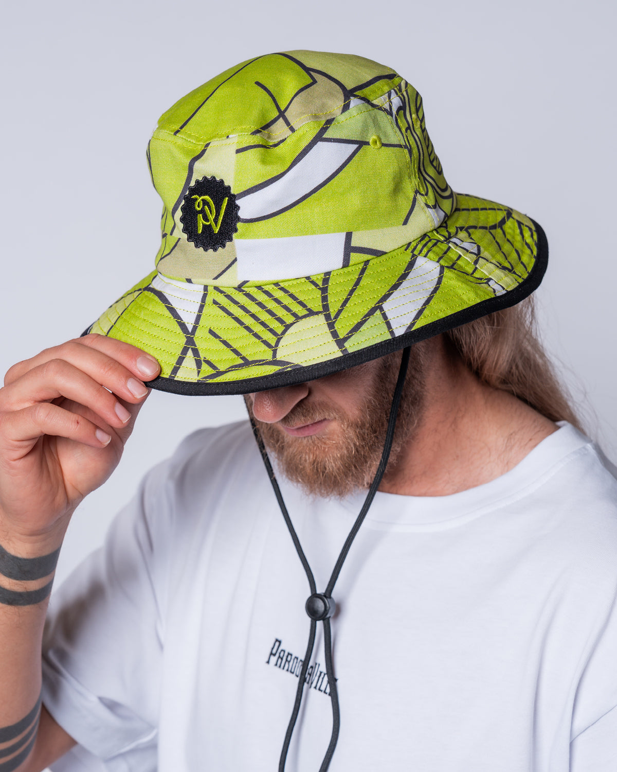 (DE) Ein Mann trägt den Safari Hat mit Bill Print. Der Hut ist gemustert in weiß und verschiedenen Grüntönen. Außerdem hat er vorne ein grünes PV Logo auf einem schwarzen Emblem. (EN) A man wears the safari hat with bill print. The hat is patterned in white and various shades of green. It also has a green PV logo on the front on a black emblem.