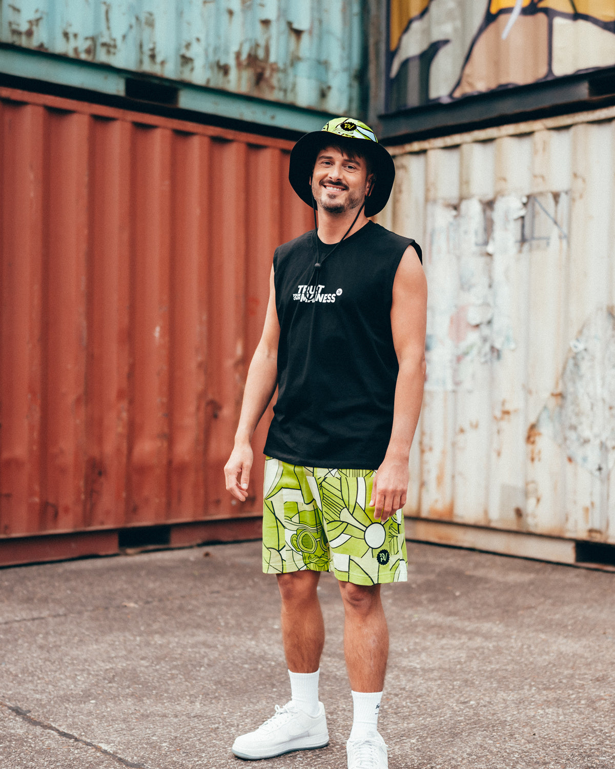 (DE) Ein Mann trägt die Shorts mit Bill Print, gemustert in hellgrün und weiß. Außerdem trägt er dazu den passenden Bucket Hat und ein schwarzes ärmelloses Shirt mit weißem Schriftzug. (EN) A man wears the shorts with Bill print, patterned in light green and white. He is also wearing the matching bucket hat and a black sleeveless shirt with white lettering.