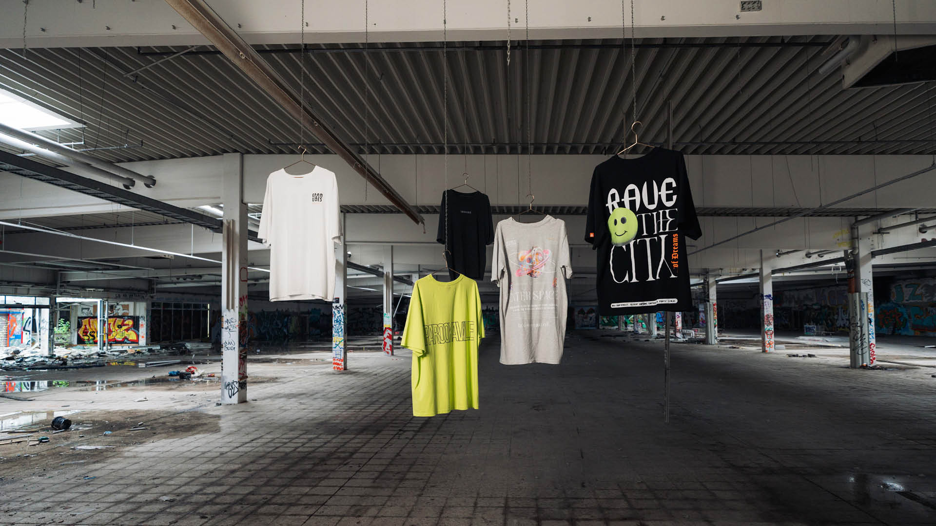(DE) Man sieht verschiedene T-Shirts aufgehängt vor einem bunten Graffiti Hintergrund in den Farben weiß, neongrün grau und schwarz. (EN) One can see different T-shirts hanging in front of a colourful graffiti background. The colours of the shirts are white, neon green, grey and black.
