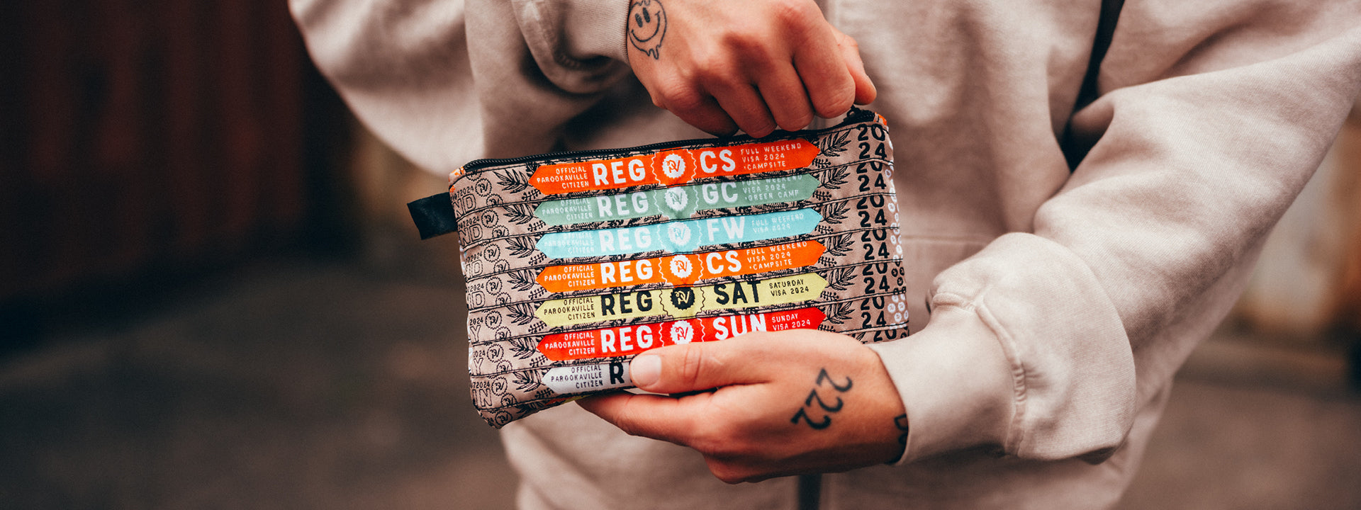 (DE) Die Kulturtasche ist braun mit schwarzer Palmenblattbestickung. Rechts ist vielfach die Jahreszahl 2024 zu sehen, in der Mitte sind verschiedenfarbige Einlassbändchen der einzelnen Camps und Visen zu sehen. (EN) The toiletry bag is brown with black palm leaf embroidery. The year 2024 can be seen in many places on the right, and in the centre there are different coloured admission ribbons for the individual camps and visas.