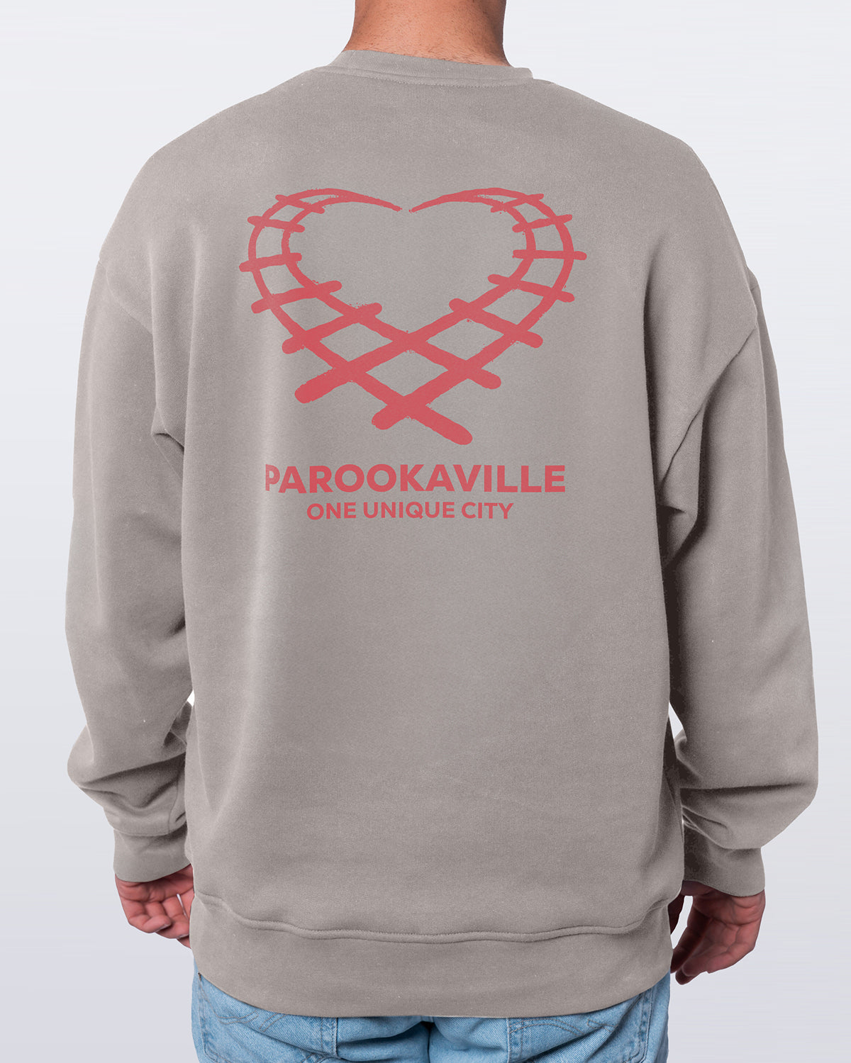 (DE) Ein Mann trägt einen hellgrauen Heart Sweater mit Backprint. Auf dem Backprint ist in Rot ein Herz aus einem Schienenmuster und darunter der Schriftzug "PAROOKAVILLE ONE UNIQUE CITY" zu sehen. (EN) A man is wearing a light grey heart jumper with a back print. The back print features a red heart with a rail pattern and the words ‘PAROOKAVILLE ONE UNIQUE CITY’ underneath.