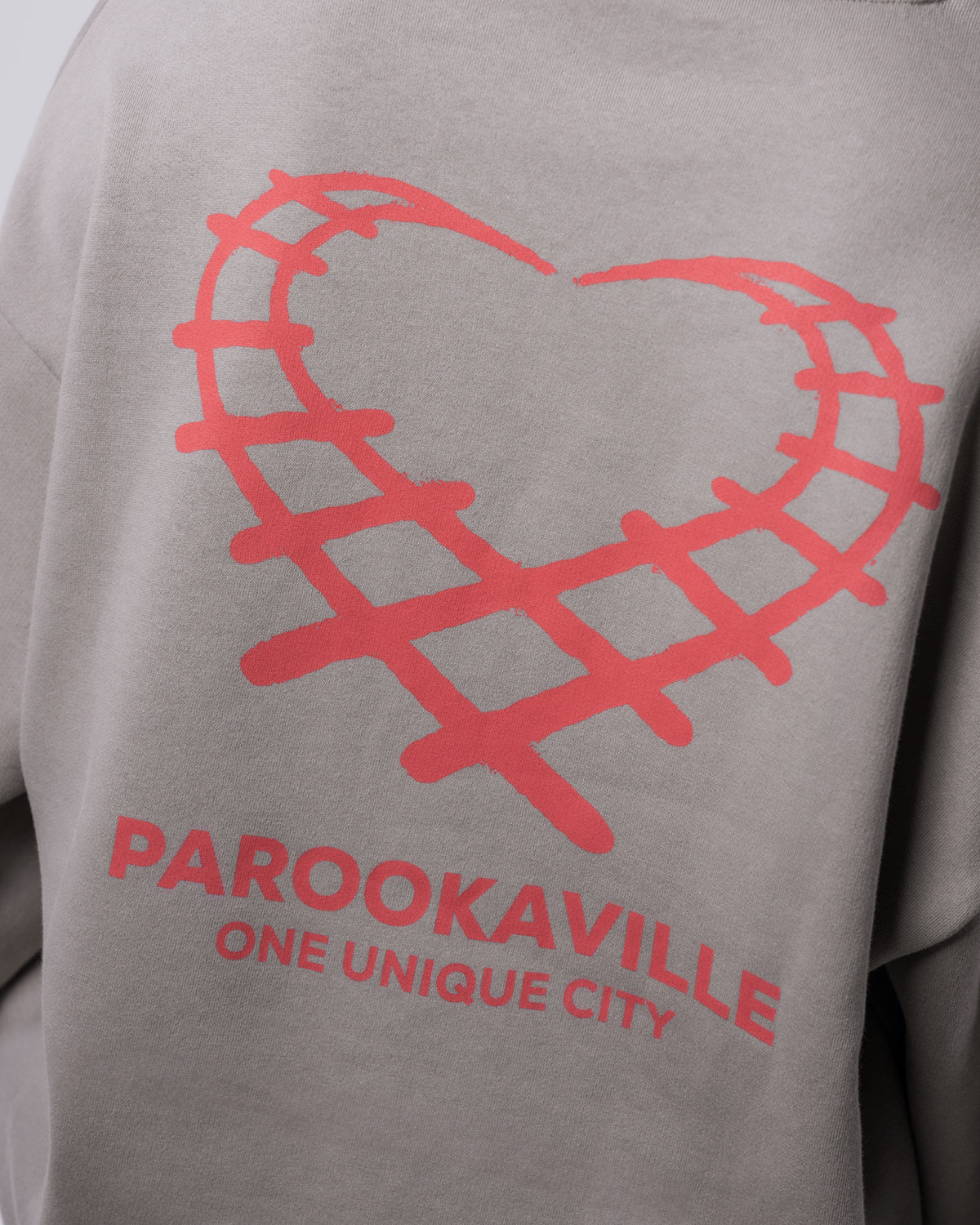 (DE) Man sieht eine Nahansicht des roten Back Prints auf dem grauen Heart Sweater. Dieser besteht aus einem Herz aus Schienenmustern und darunter ist der Schriftzug "PAROOKAVILLE ONE UNIQUE CITY". (EN) One can see a close-up view of the red backprint of the grey Heart sweater. It is heart shaped with a rail pattern and underneath is the inscription "PAROOKAVILLE ONE UNIQUE CITY".