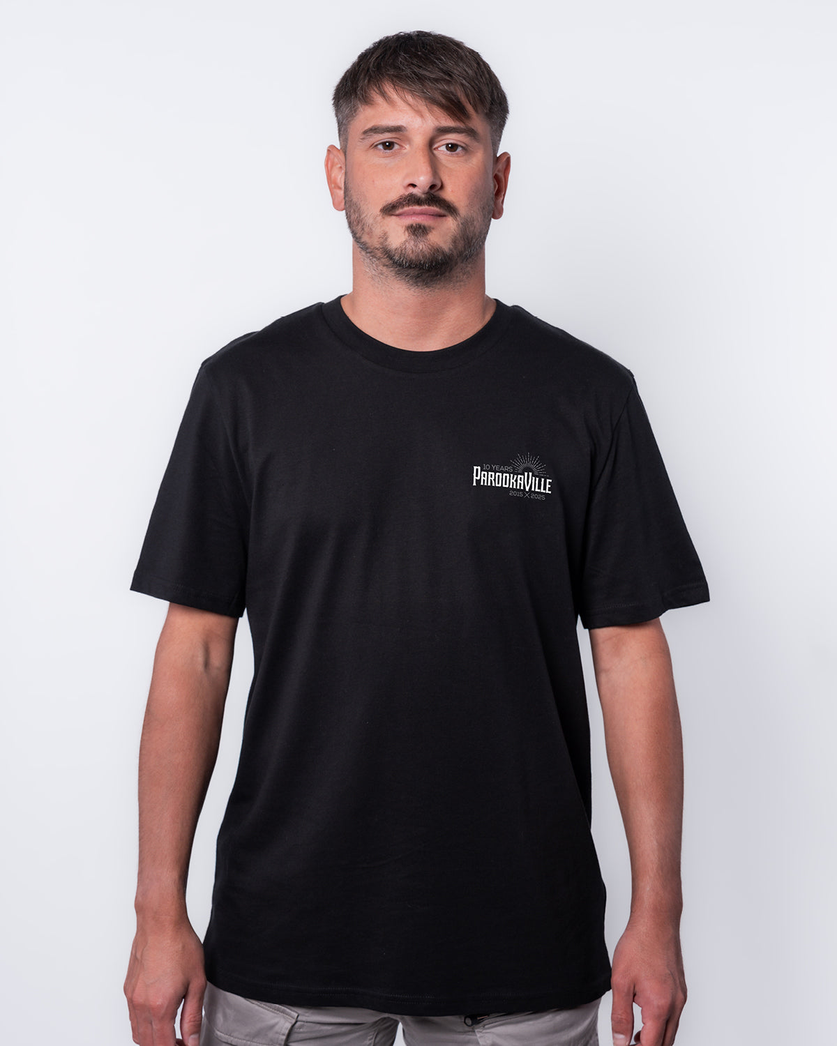 (DE) Ein Mann trägt ein schwarzes T-Shirt mit dem weißen 10 Years PAROOKAVILLE Schriftzug auf der rechten Oberseite. (EN) A man is wearing a black T-shirt with the white 10 Years PAROOKAVILLE lettering on the top right.