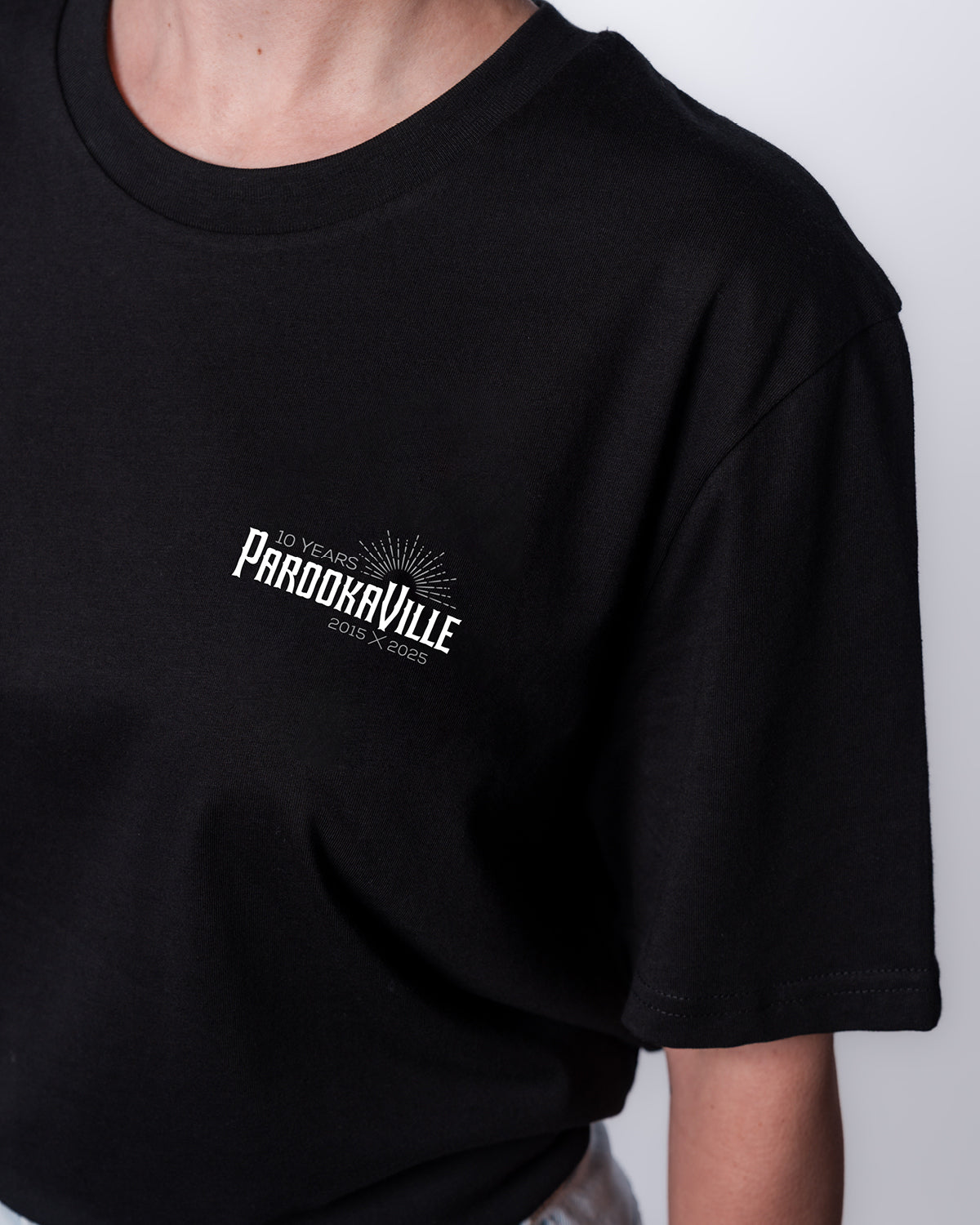 (DE) Man sieht eine Nahansicht eines schwarzes T-Shirts mit dem weißen 10 Years PAROOKAVILLE Schriftzug auf der rechten Oberseite. (EN) You can see a close-up view of a black T-shirt with the white 10 Years PAROOKAVILLE lettering on the top right.