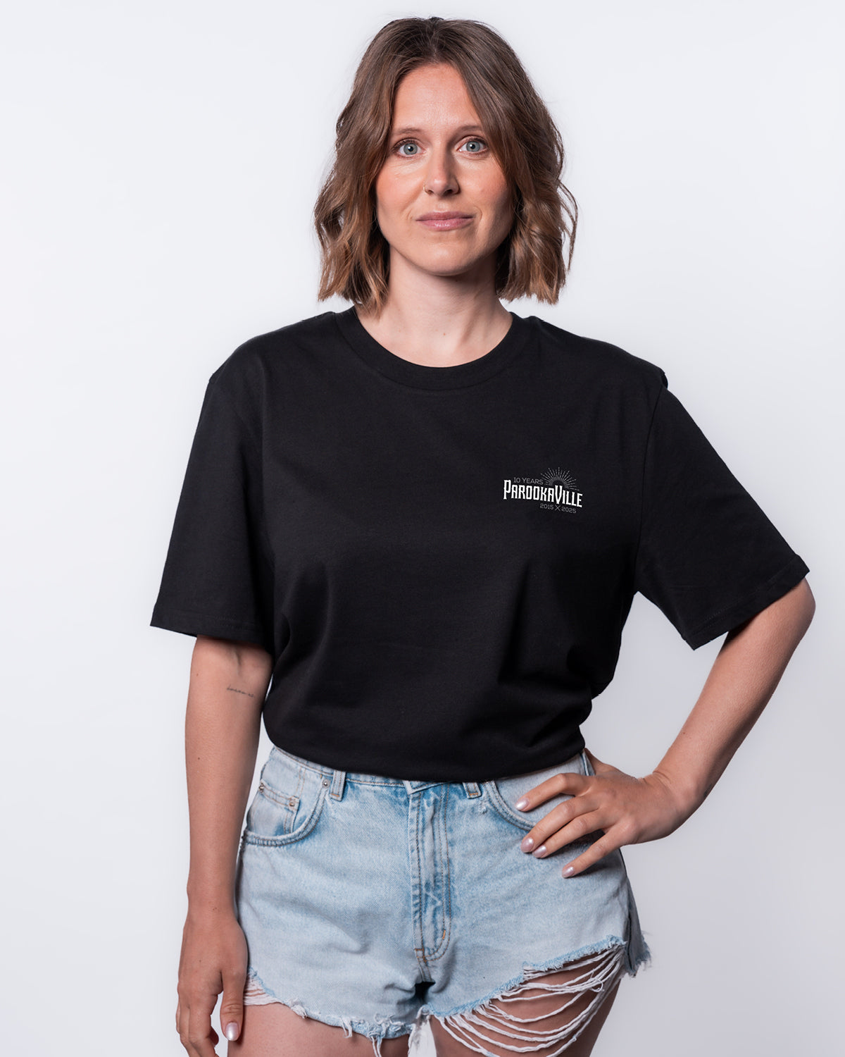 (DE) Eine Frau trägt ein schwarzes T-Shirt mit einem weißen 10 Years PAROOKAVILLE Schriftzug. Dazu trägt sie eine kurze, ausgefranste Jeans Shorts. (EN) A woman is wearing a black T-shirt with white 10 Years PAROOKAVILLE lettering. She is also wearing short, frayed denim shorts.