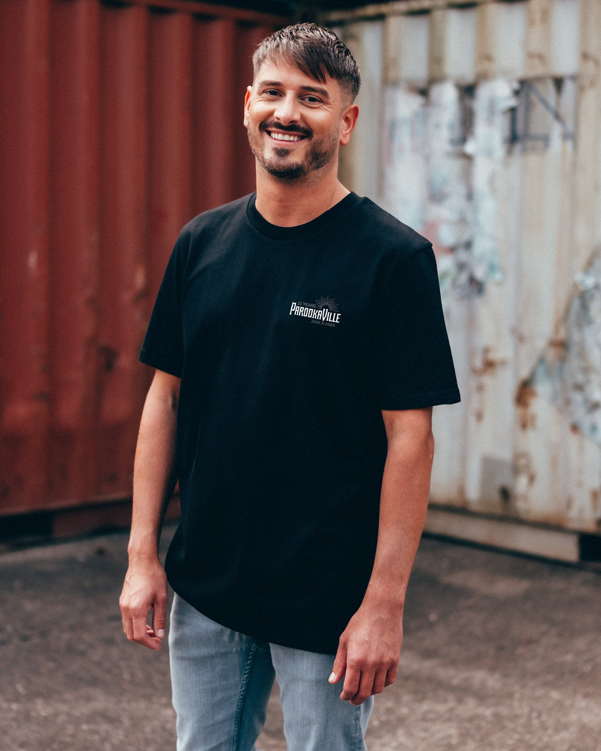 (DE) Ein Mann steht vor bunten Containern. Er trägt ein schwarzes T-Shirt mit weißem 10 Years PAROOKAVILLE-Logo auf der linken Oberseite. (EN) A man stands in front of colourful containers. He is wearing a black T-shirt with a white 10 Years PAROOKAVILLE logo on the top left.