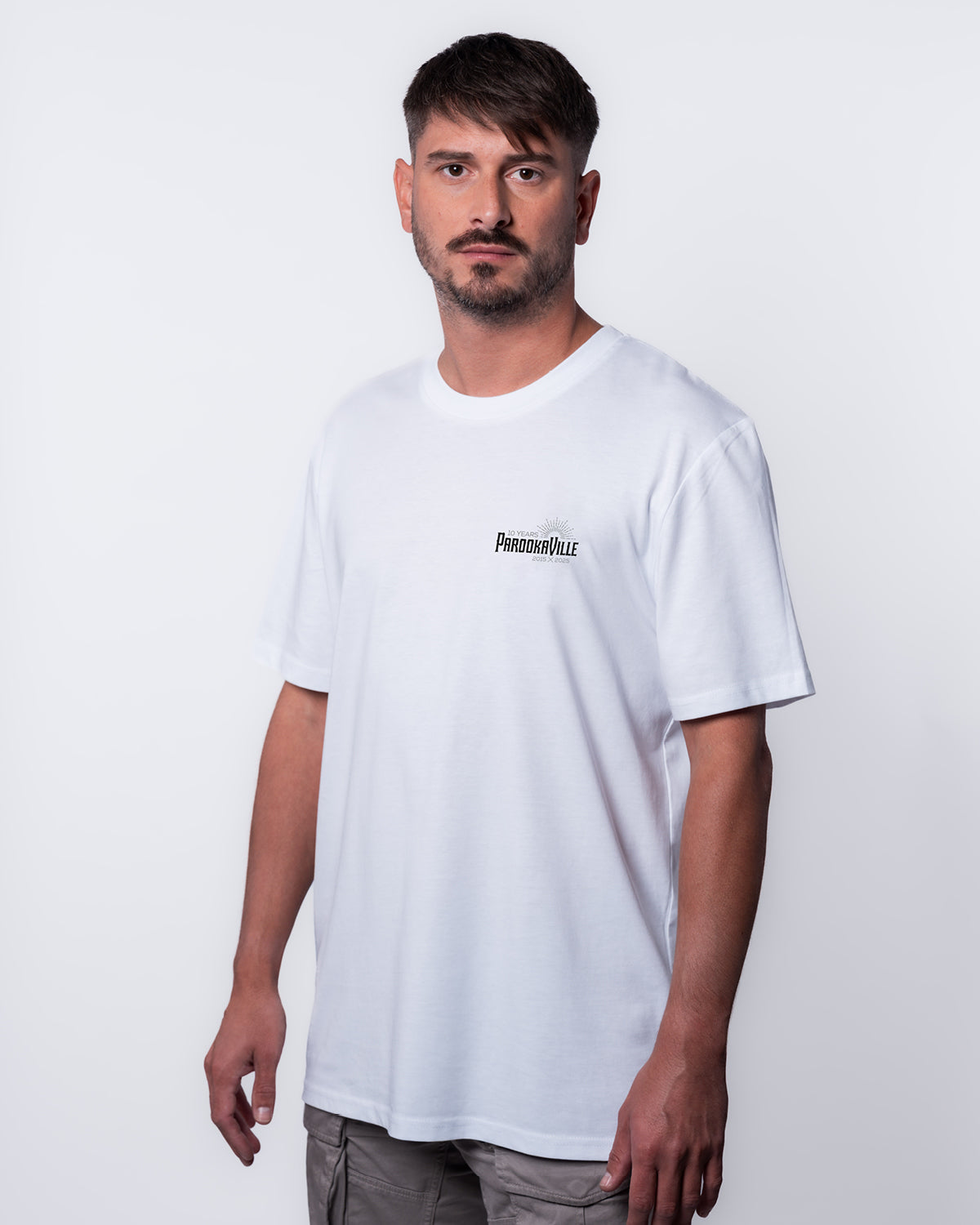 (DE) Ein Mann trägt ein weißes T-Shirt mit dem schwarzen 10 Years PAROOKAVILLE Schriftzug. Das T-Shirt fällt locker am Körper und sieht bequem aus. (EN) A man wears a white T-shirt with the black 10 Years PAROOKAVILLE lettering. The T-shirt falls loosely on the body and looks comfortable.