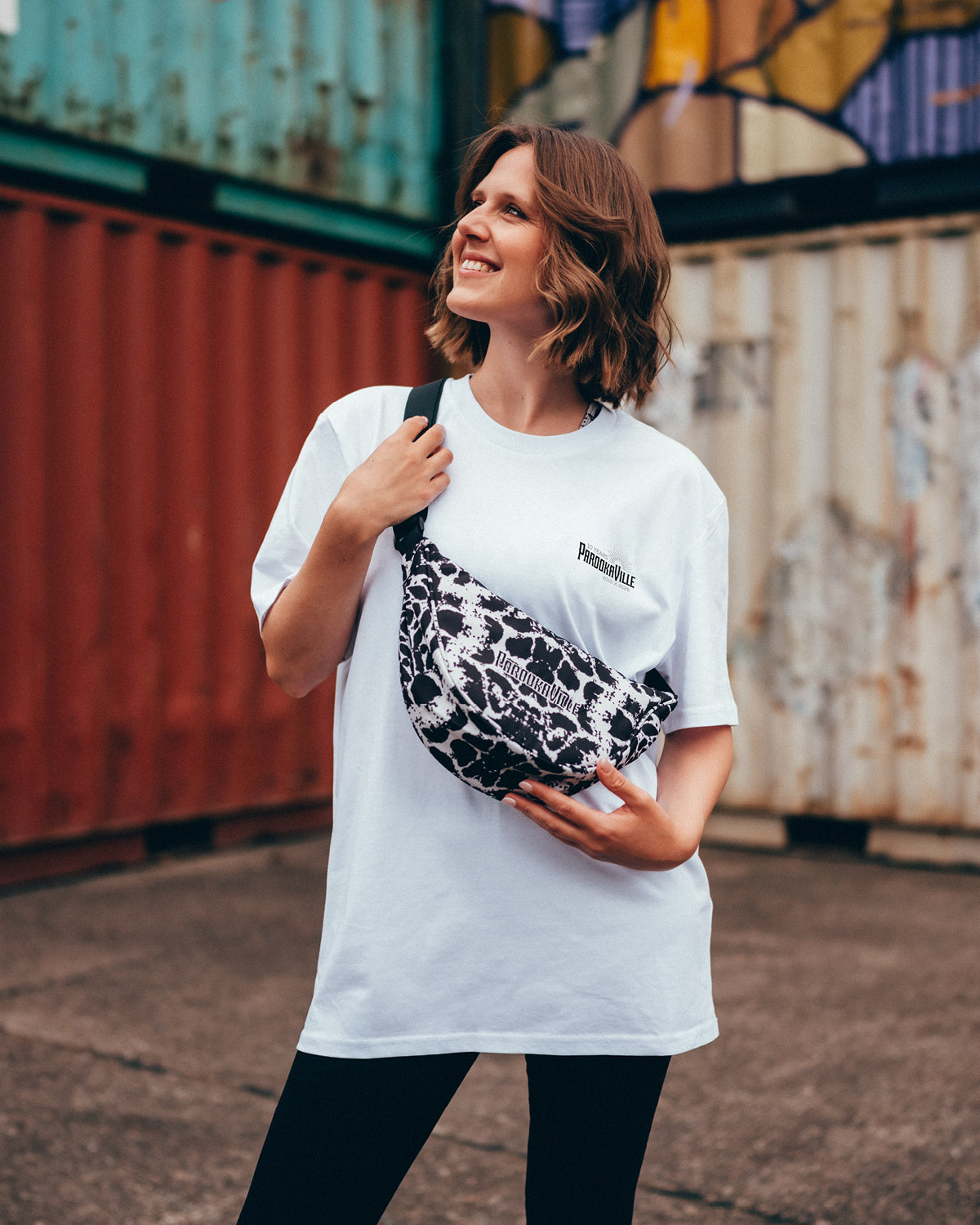 (DE) Eine Frau steht vor bunten Containern. Sie trägt die schwarz-weiß gefleckte Moonbag mit Leopardenmuster und dazu ein weißes T-Shirt mit schwarzem PAROOKAVILLE Schriftzug oben rechts. (EN) A woman stands in front of colourful containers. She is wearing a black and white spotted moonbag with a leopard print and a white T-shirt with black PAROOKAVILLE lettering on the top right.