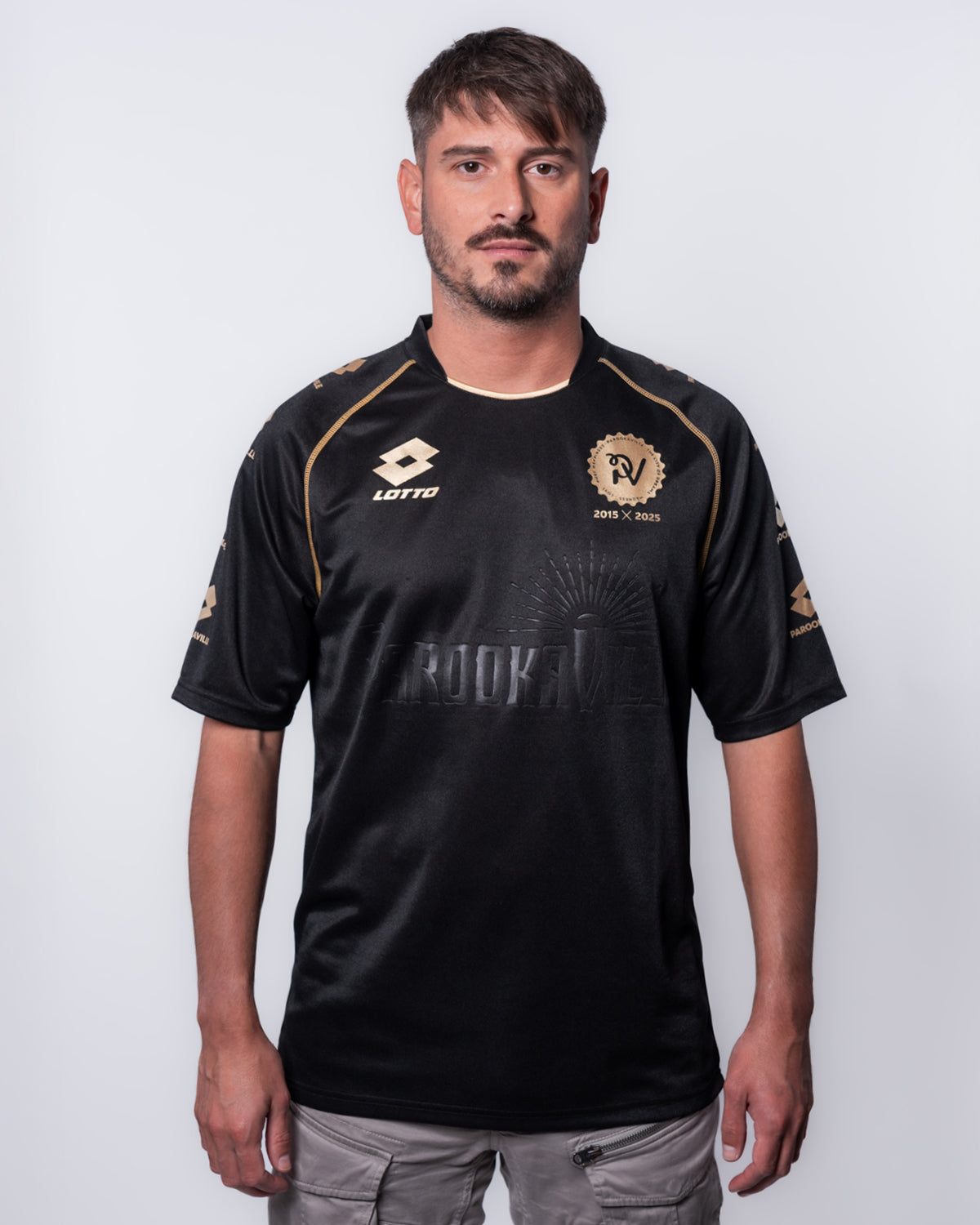 (DE) Ein Mann trägt ein schwarzes 10 Years PAROOKAVILLE Trikot. Vorne sieht man auf der linken Oberseite das Lotto- Emblem und auf der rechten Seite das PV 2015X2025 Emblem, jeweils in Gold. (EN) A man is wearing a black 10 Years PAROOKAVILLE jersey. On the front you can see the Lotto emblem on the top left and the PV 2015X2025 emblem on the right, both in gold.