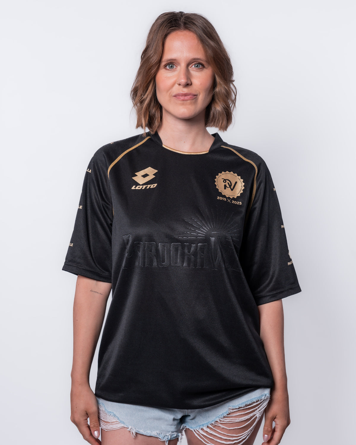 (DE) Eine Frau trägt das schwarze 10 Years PAROOKAVILLE Trikot. Vorne sieht man oben links das Lotto-Emblem, rechts das PV 2015X2025 Emblem jeweils in Gold. (EN) A woman wears the black 10 Years PAROOKAVILLE jersey. The Lotto emblem can be seen at the top left of the front and the PV 2015X2025 emblem in gold on the right.