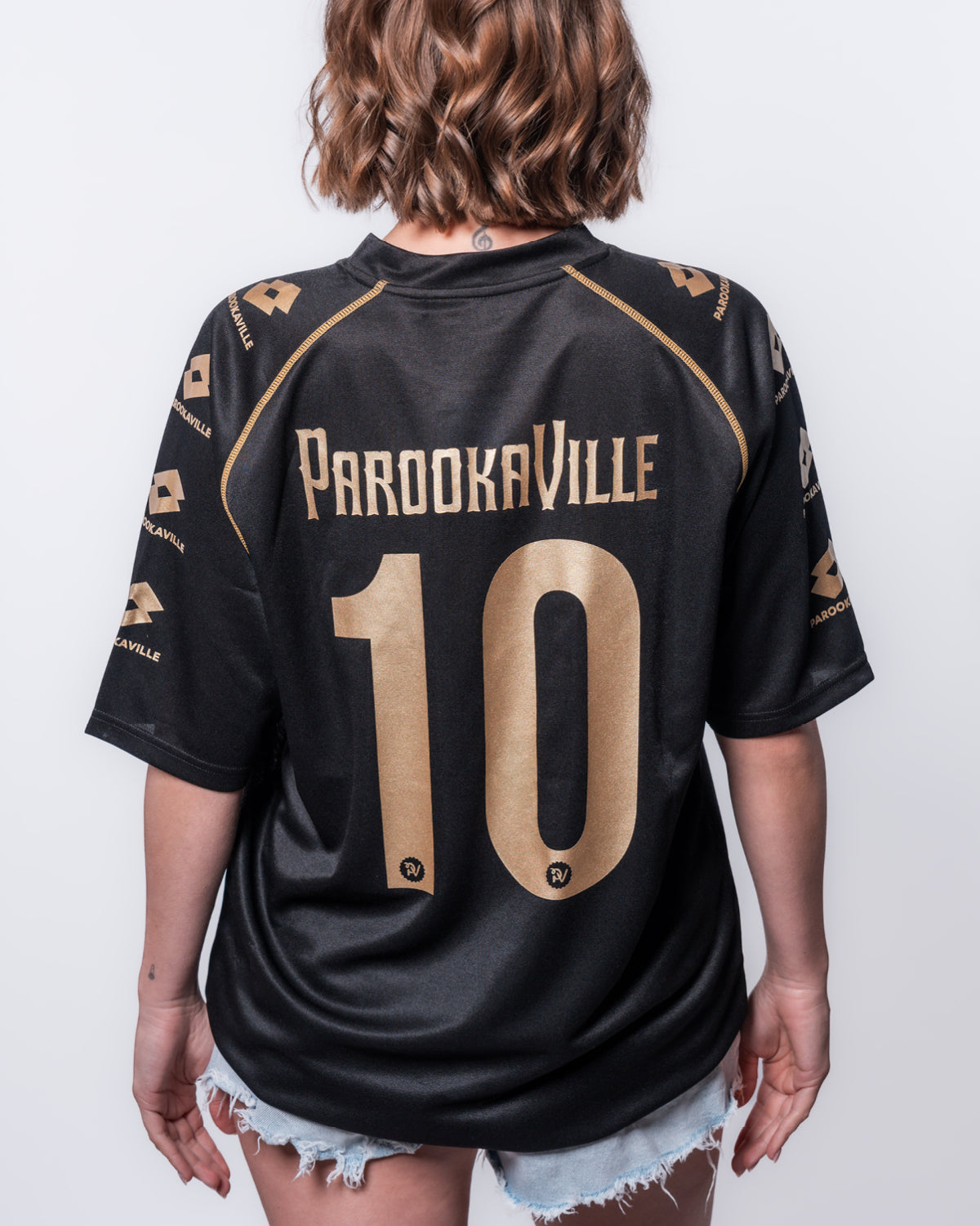 (DE) Eine Frau trägt das schwarze PAROOKAVILLE 10 Years Trikot. Man sieht sehr gut den Backprint "PAROOKAVILLE 10" in goldener Schrift. (EN) A woman wears the black PAROOKAVILLE 10 Years jersey. You can clearly see the back print ‘PAROOKAVILLE 10’ in gold lettering.